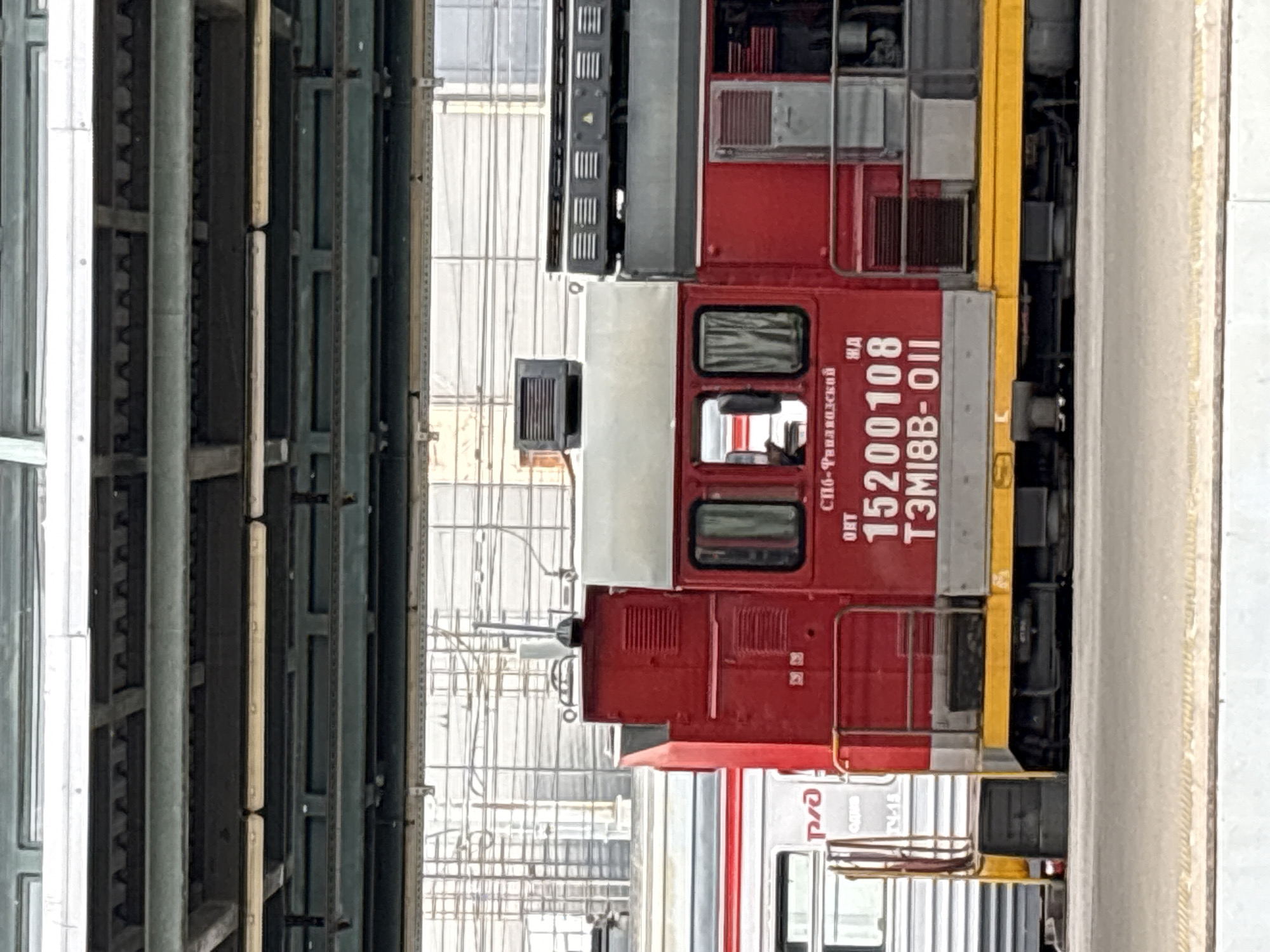 A red diesel locomotive is parked at a train station in Russia. The locomotive is primarily red with a yellow stripe along the bottom. It has a cab with two windows, curtains visible inside. The number "15200108" is painted on the side of the locomotive, along with "ТЭМІВВ-ОІІ" below it. Text above the number reads "СПБ - Финляндский". The locomotive is positioned next to another train car, which is mostly silver with red trim and appears to have the letters "Р/Э" on it. The scene is viewed from the side, with a portion of a covered walkway or bridge visible overhead. The background shows a complex structure of wires and poles, indicative of a railway infrastructure. The ground in the foreground is a light-colored surface, possibly a platform.
