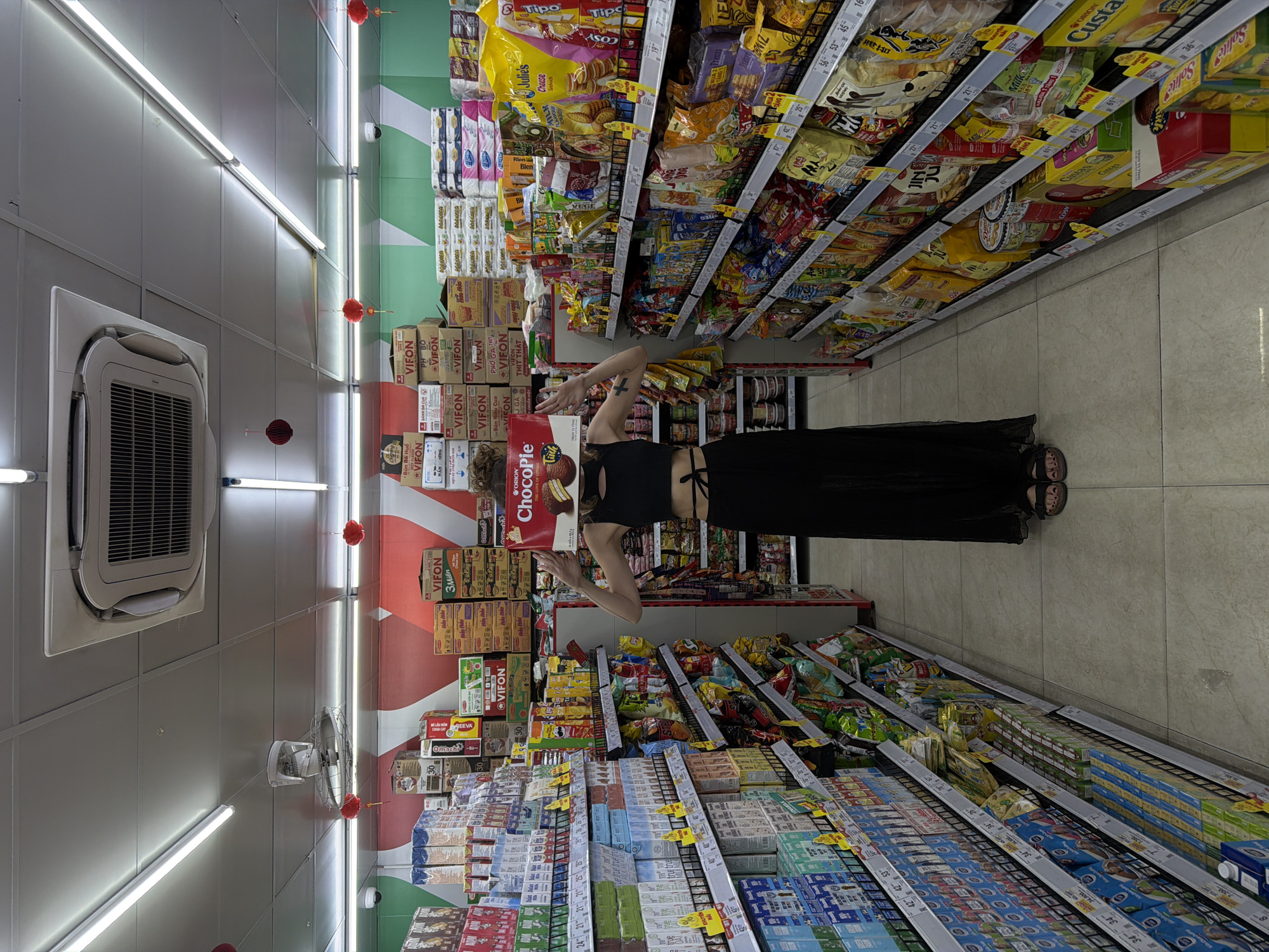 The image displays a person posing in a brightly lit supermarket aisle in Đà Nẵng, Vietnam. The individual, wearing a black crop top and a long black skirt with sandals, stands with a large red "Orion ChocoPie" box held in front of their face, obscuring it. Their arms are raised, cradling the box, suggesting a playful or staged photo.
The aisle is narrow, with shelves packed full of various packaged food products on both sides. Prominent Vietnamese brands are visible, such as "VIFON," "Hảo Hảo," "Omachi" (instant noodles), along with snack brands like "Tipo," "Julie's," "Cosy," "Custas," and "Solite." International brand "Milka" is also present. Text on packaging includes "TRỌN VỊ TÌNH," "Bún Riêu Cua," and "Phở Gà Thịt Thật." Prices are displayed on shelf labels.
The ceiling features multiple fluorescent light fixtures, and red spherical lanterns are hung periodically. An air conditioning unit and a wall-mounted fan are visible on the left wall. Cardboard boxes, many bearing the "VIFON" logo, are stacked on the floor and behind the shelves, indicating inventory. The floor is tiled. The scene suggests a typical, well-stocked grocery store environment in Vietnam.