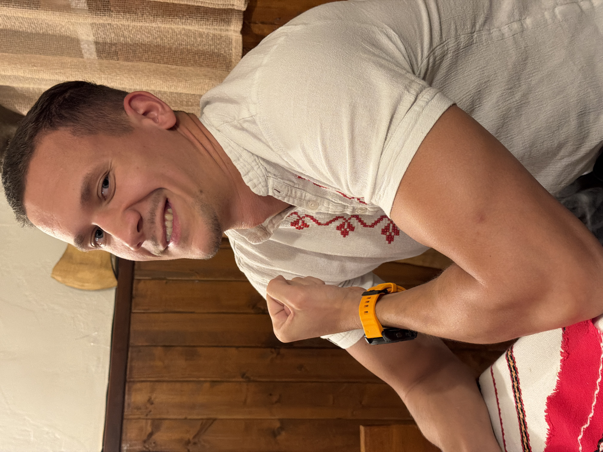 A man is posing for a photograph in an indoor setting, likely within a traditional Bulgarian-themed establishment. He has fair skin, short brown hair, and is wearing a white shirt with red embroidered details around the neckline and placket, an ornamental design typical of Bulgarian folk attire. A bright orange wristwatch with black accents is visible on his left wrist. The man is smiling and appears to be facing the camera at an angle.
The backdrop includes a wooden paneling, potentially wall or furniture, and patterned fabric that appears to be part of the setting's decor. The photograph is taken at an angle, giving a partial view of what seems to be a pillow or cushion with red, white, and black stripes.
The location is Gabrovo, Bulgaria, as indicated by the traditional attire and the potential ambiance.