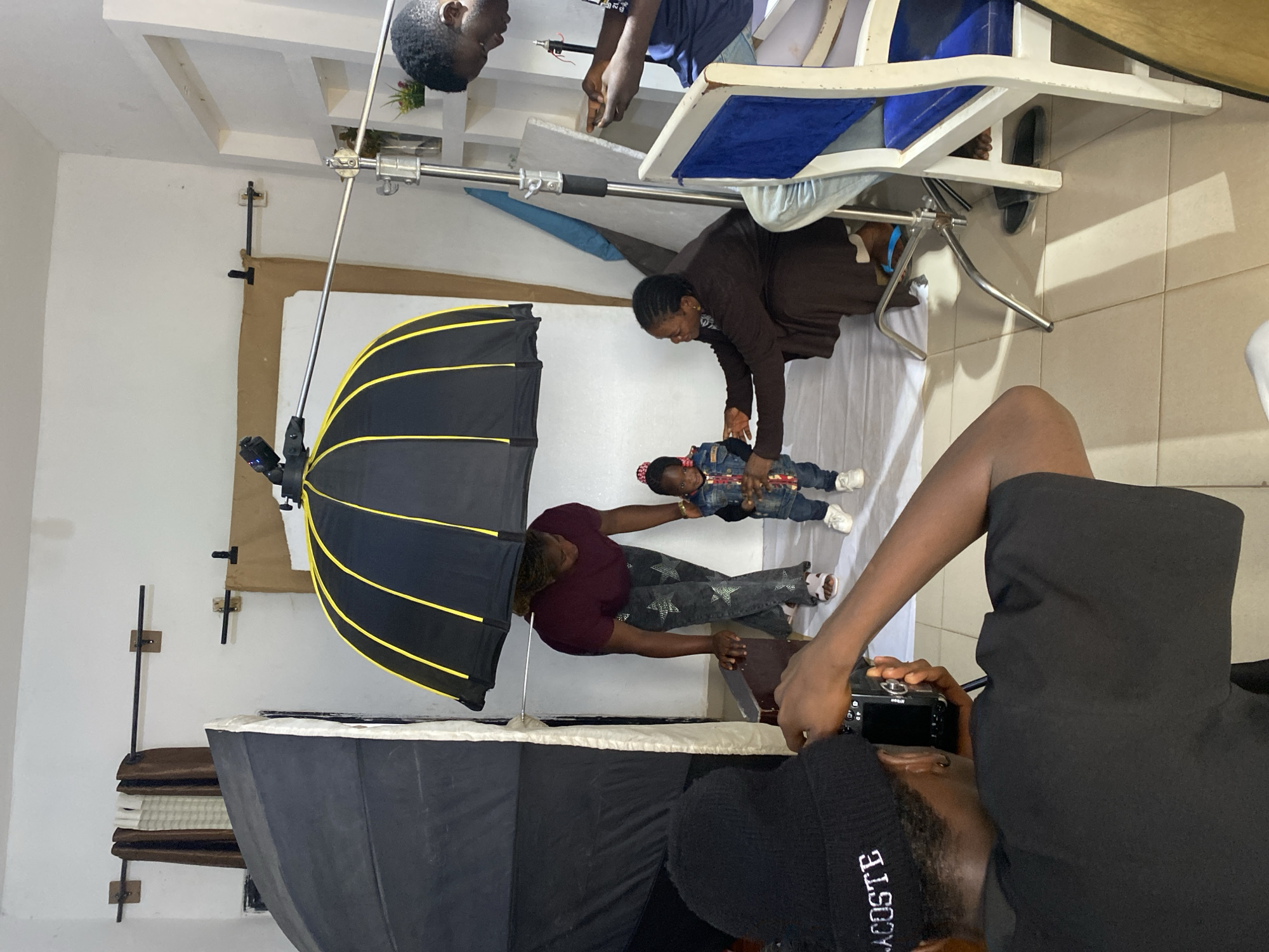 In a studio setting in Bukuru, Nigeria, a photographer wearing a black beanie and a black t-shirt is positioned in the foreground, holding a camera and actively taking a photograph. To the right of the photographer, a young child is standing and being attended to by two adults. One adult, wearing a maroon shirt and patterned pants, stands behind the child, seemingly supervising. Another adult, in a brown outfit, is crouched down in front of the child, adjusting their clothing or pose. A large umbrella-shaped lighting fixture with yellow accents is positioned above the child, casting light on the scene. The studio appears to be set up with a white backdrop, and various studio equipment, including light stands and a large softbox, are visible. The overall impression is a professional or semi-professional photoshoot in progress.