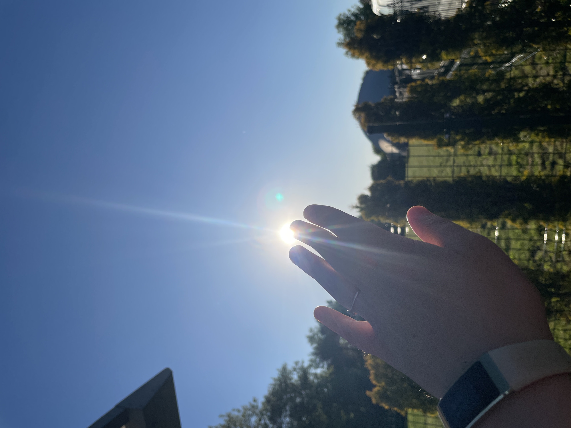 A low-angle shot captures a hand held towards the sun in a clear blue sky. The sun is positioned between the fingers, causing a bright flare. The hand has a silver ring on one finger and a fitness tracker on the wrist. Background elements include trees and a building with a triangular roof, with a fence visible amongst the trees. The location is potentially a park or green space in an urban setting.