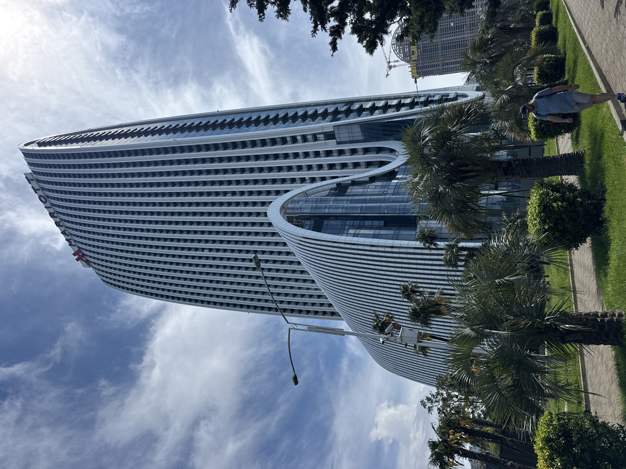 The image presents a tall, modern skyscraper with a curved facade, located in Batumi, Georgia. The building's exterior is characterized by horizontal white lines and glass panels. A portion of the structure has a curved architectural design. Palm trees and greenery are present at the base of the building. A pedestrian walkway borders the greenery. A man is walking on the walkway. A lamppost stands adjacent to the building. In the background, another building is under construction, with visible scaffolding and cranes. The sky is blue with scattered clouds.