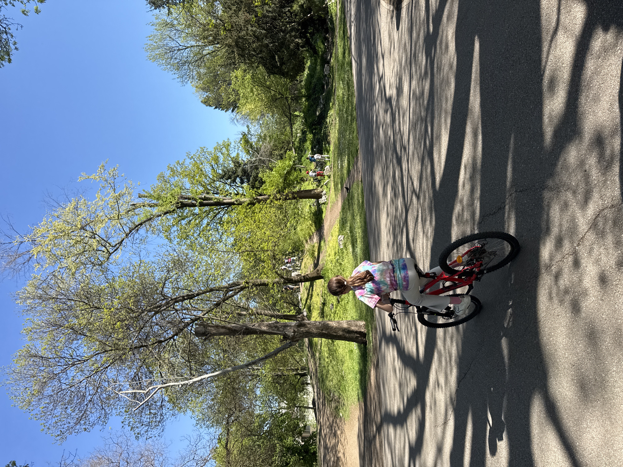 A young girl rides a red bicycle on an asphalt path in a park. The girl has long brown hair tied back in a ponytail and wears a colorful tie-dye shirt, light-colored pants, and white shoes. The path is lined with trees that cast shadows across the pavement. In the background, there is green grass and more trees, with a few people visible in the distance. The sky is blue and clear. Based on the trees, and the style of park, this is most likely in the city of Sofia, Bulgaria.
