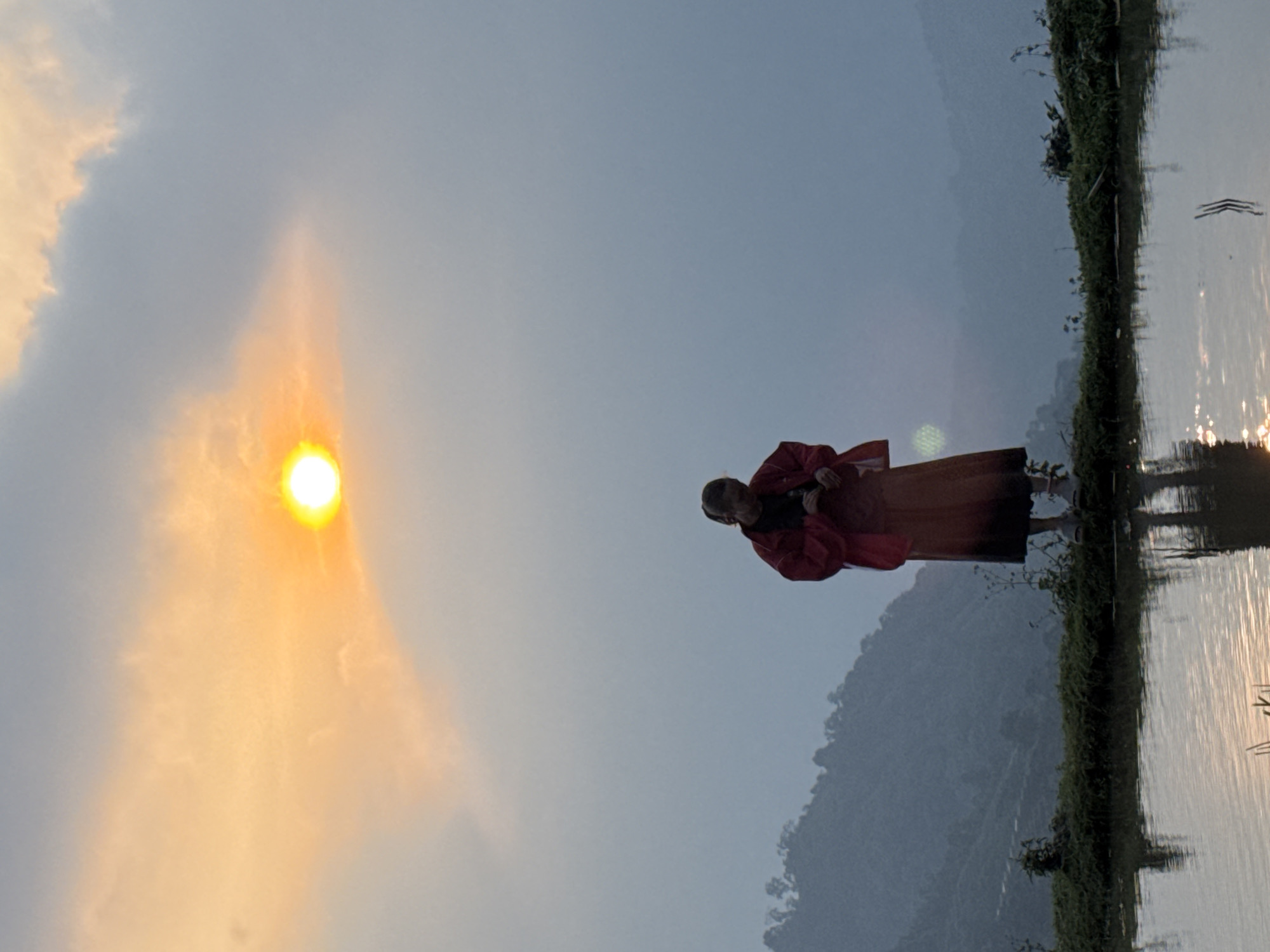 The image depicts an outdoor scene at either sunrise or sunset in Nậm Dịch Commune, Vietnam. A single person stands in calm water, facing towards the left where a bright, glowing sun is visible in the hazy sky.
**Subjects:** The main subject is a person, likely an adult, dressed in a red top or jacket and a dark skirt or lower garment. Their silhouette and partial reflection are visible in the water. The surrounding environment includes the sun, clouds, water, distant mountains, and green vegetation along the water's edge.
**Scene/Setting:** The setting is a body of water, possibly a rice paddy field, lake, or river, bordered by a strip of green foliage on the right side. In the background, dark, hazy mountains rise under a light blue-grey sky.
**Activity or Event:** The person appears to be standing contemplatively in the water, looking towards the sun. Their posture suggests a moment of appreciation for the natural beauty.
**Notable details:**
* **Time of day:** The prominent, bright orange sun low in the sky, casting a warm glow and light flares, strongly indicates either early morning (sunrise) or late evening (sunset). The sky is a blend of soft orange near the sun and light blue-grey further away, suggesting a transition period of light.
* **Weather:** The sky is partially cloudy and somewhat hazy, contributing to the diffused light around the sun.
* **Location cues:** The presence of mountains, water, and vegetation is consistent with rural, possibly agricultural, landscapes found in regions like Nậm Dịch Commune, Vietnam. The water surface is still enough to provide clear reflections of the person and the sky, with some ripples from the sun's glare.
* **Emotions:** While not explicitly clear, the scene evokes a sense of peace and tranquility, possibly contemplation or enjoyment of nature by the person.
**Visible text:** There is no visible text in the image.