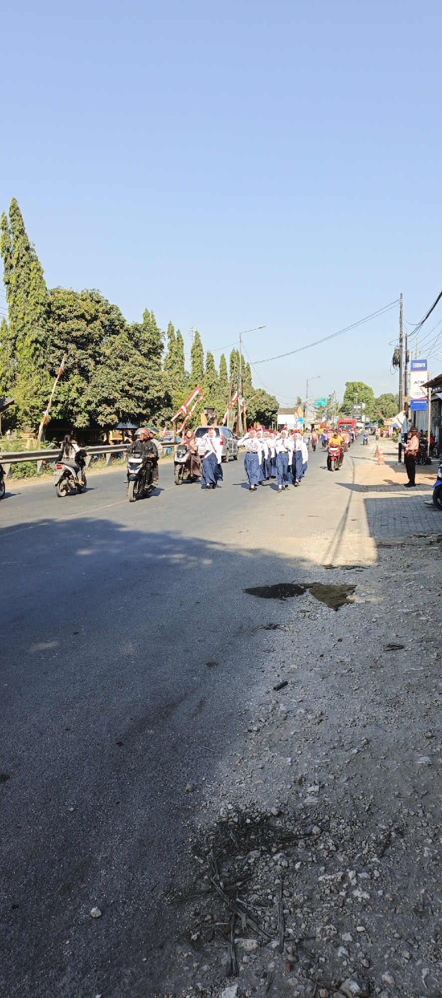 In Kasiyan Timur, Indonesia, on a bright, clear day, a procession of uniformed students walks down a paved road. The students, appearing to be male and female, are dressed in white shirts, red headscarves, and blue pants or skirts, suggesting a school uniform. They march in formation, with some carrying red and white flags. Several motorbikes are present on the road, with one rider stopped on the side. A small, light-colored car and a red truck are also visible in the background. The roadside features trees, some tall and slender, others more full, and various buildings, including one with an ATM sign. A railroad crossing gate is visible on the left side of the road, and traffic signals and utility poles line the street. The road surface shows signs of wear, with some patches of gravel and dark stains, possibly from oil or water. The overall atmosphere suggests a community event or a parade.