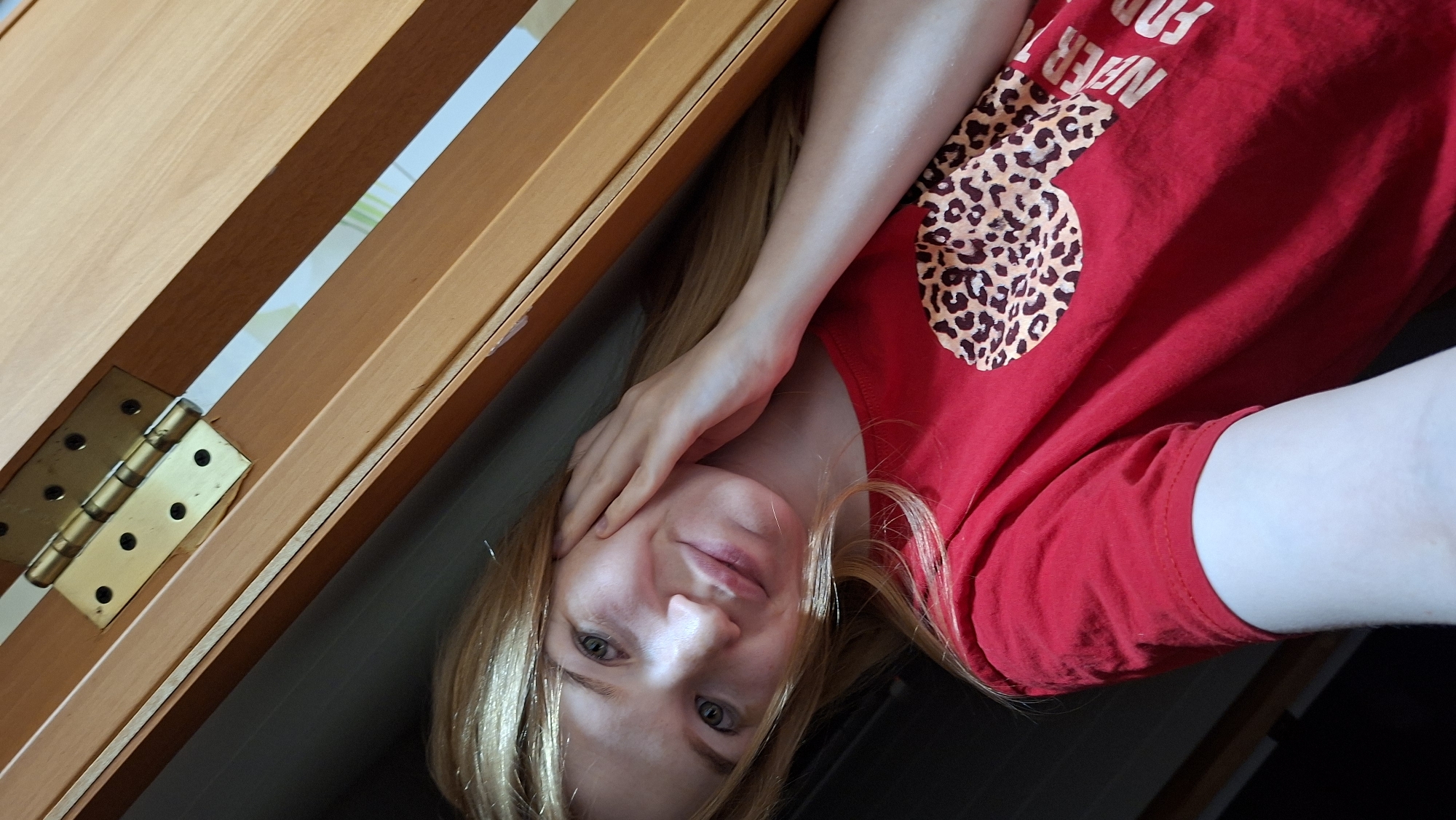 A young Caucasian female with long blonde hair and light blue eyes is taking a selfie. She is wearing a red t-shirt with a leopard print design and some text. The woman is leaning against a wooden door frame, with a brass hinge visible at the top of the frame. The background is dark and out of focus. The lighting suggests an indoor environment, possibly a bedroom or closet. The scene is set in Большой Букор, Russia.