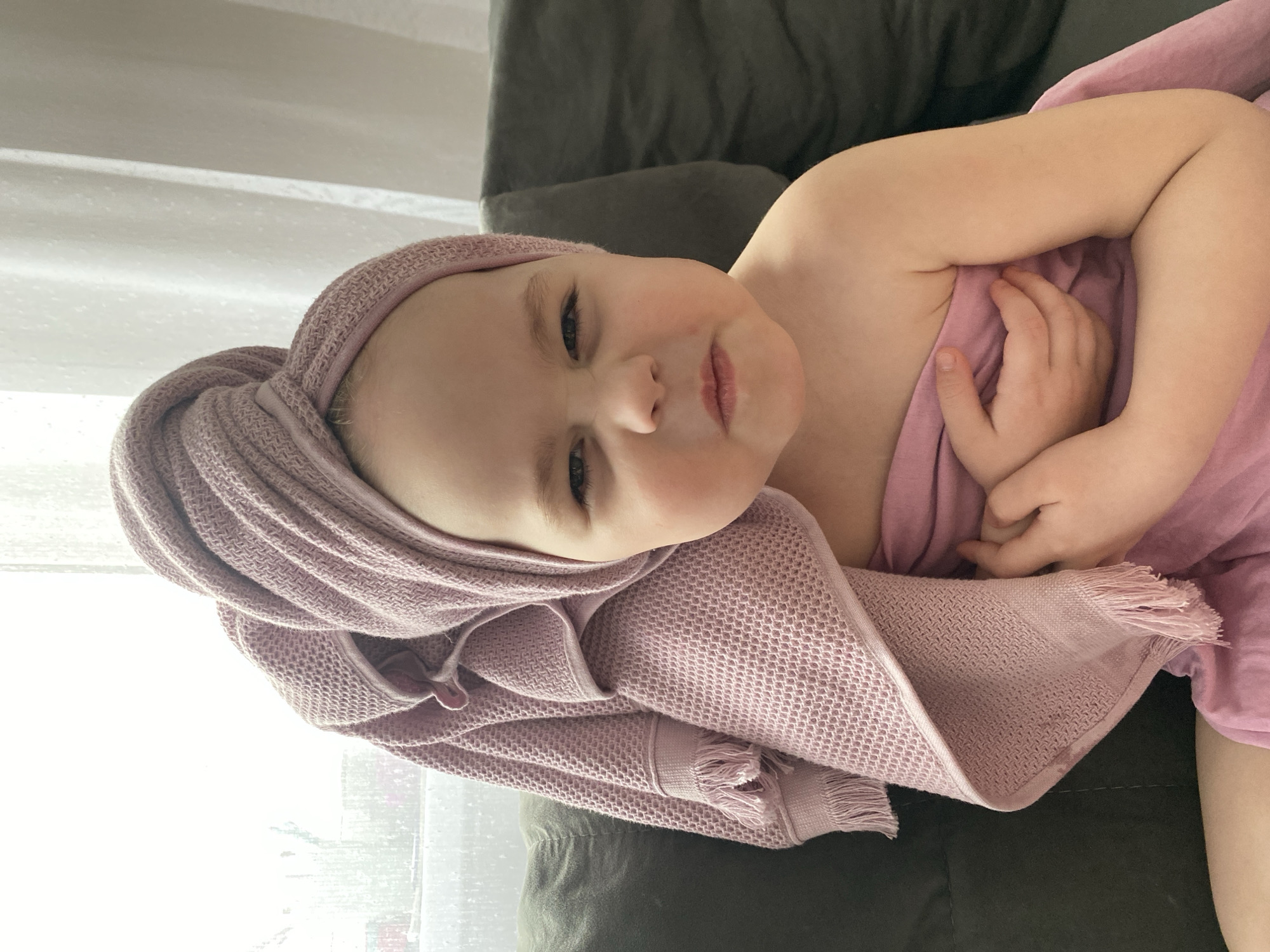 A young child, wrapped in a pink towel with a portion of it fashioned into a turban on their head, is lying on a dark grey upholstered sofa. The child is fair-skinned and is positioned close to a window covered by sheer white curtains. The scene takes place indoors during the daytime. No further information is provided to suggest the location is in Izhevsk, Russia.