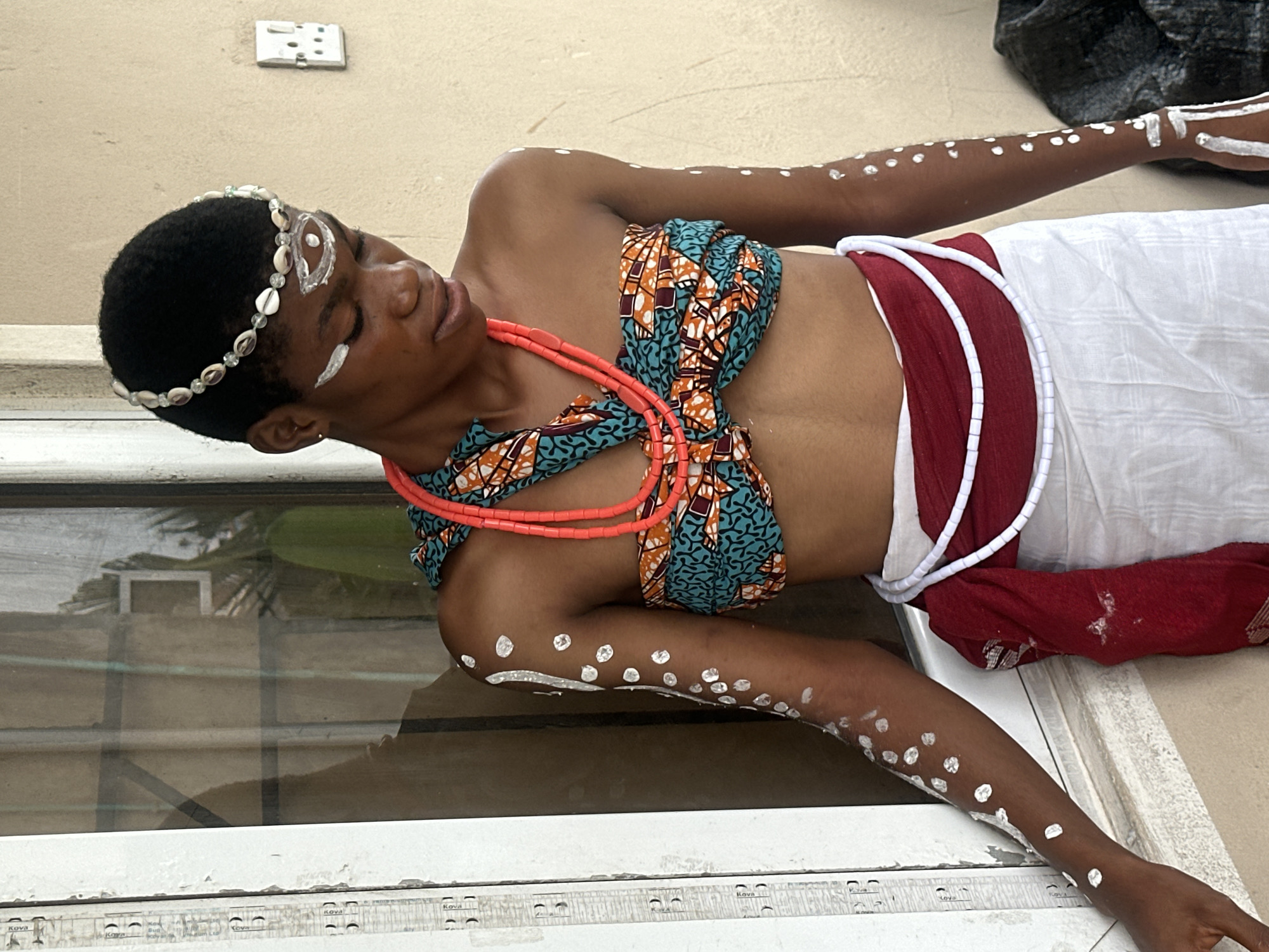 The image depicts a person adorned in traditional attire, possibly for a cultural ceremony. The subject is lying down, with their eyes closed, suggesting a state of peace or performance. They wear a beaded headdress with seashells and a patterned fabric top, complemented by coral necklaces. White markings, likely body paint, are visible on their arms, legs, and face, adding to the ceremonial aesthetic. A red fabric is wrapped around their waist, and a white cloth is draped over their legs. The background shows a textured wall and a window with reflections, indicating an outdoor or semi-outdoor setting. The presence of an electrical socket on the wall suggests a modern environment within Badagry, Nigeria. The lighting appears to be natural, implying daytime.