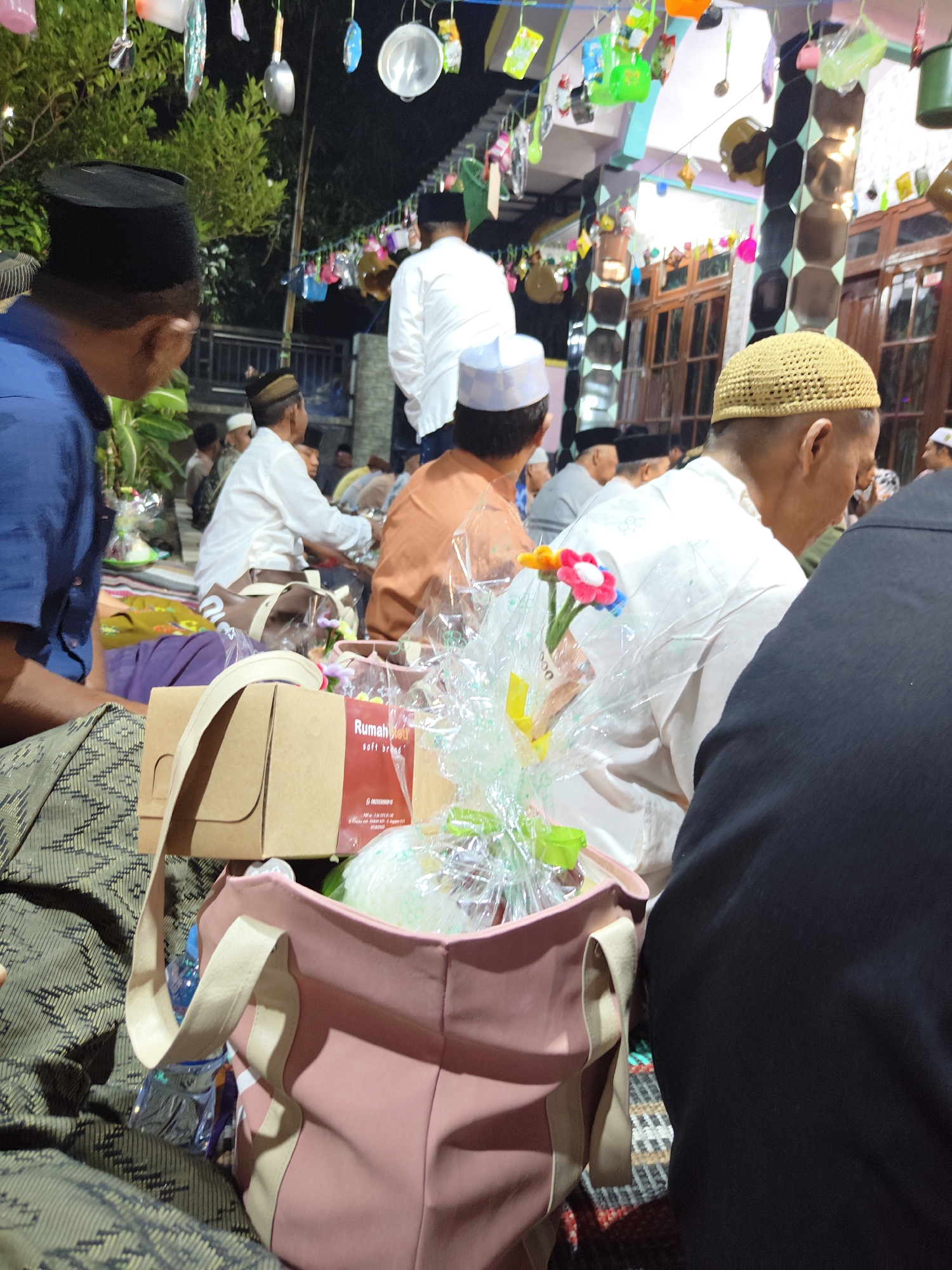 The image shows a nighttime gathering, likely an outdoor event in Cermee, Indonesia, given the presence of traditional Indonesian attire and decorations. Several individuals are present, some seated and others standing, all appearing to be men. They are dressed in traditional Islamic clothing, including peci (caps) and sarongs. In the foreground, there are gifts, including a pink tote bag with a light-colored strap, containing a water bottle and other items. Next to it are two cardboard boxes, one with a red label that reads "Rumah Bolu soft bread". There are also cellophane-wrapped items, possibly food or gifts, decorated with small artificial flowers. In the background, string lights and colorful paper decorations are visible, hanging from wires and structures. The overall atmosphere appears to be festive and communal.