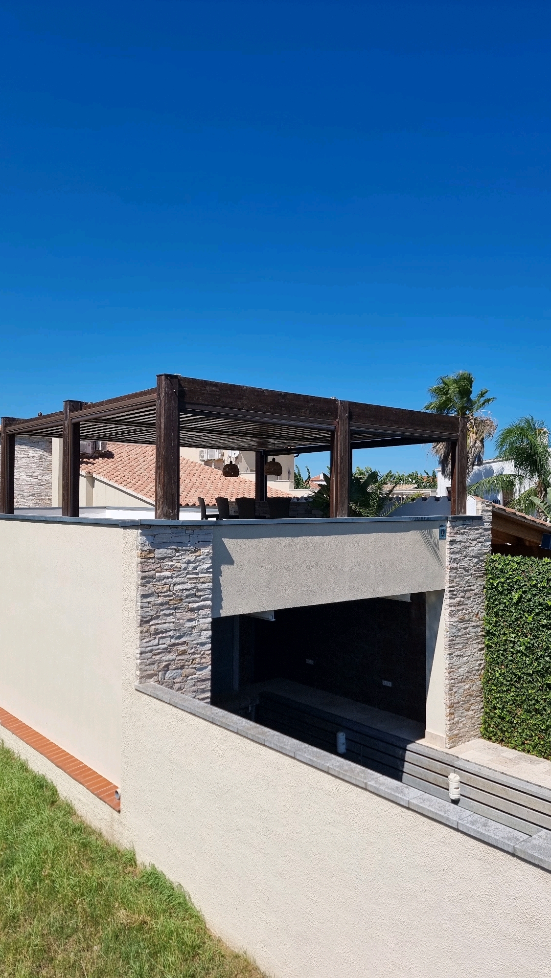 The image depicts an outdoor scene, likely a rooftop or elevated patio, under a clear blue sky. The primary subject is a dark wooden pergola, supported by thick columns, covering a seating area with chairs and a table. The pergola's roof is made of parallel wooden slats.
Below the pergola, there is a section of roof with terracotta tiles. The surrounding structure features a mix of materials: a cream-colored stucco wall, sections of stacked stone veneer in earthy tones, and a vibrant green hedge on the right side of the frame. A grassy slope with a small red brick border is visible in the lower left corner.
The scene appears to be residential, possibly a villa or a building in a resort area. The presence of palm trees in the background suggests a warm climate. The lighting indicates it is daytime, with bright sunlight casting shadows. No people are visible in the image.
While the image is located in Castelló d'Empúries, Spain, there are no specific landmarks or clues within the visual data to pinpoint an exact location within the city. The scene presents a well-maintained, modern outdoor living space.