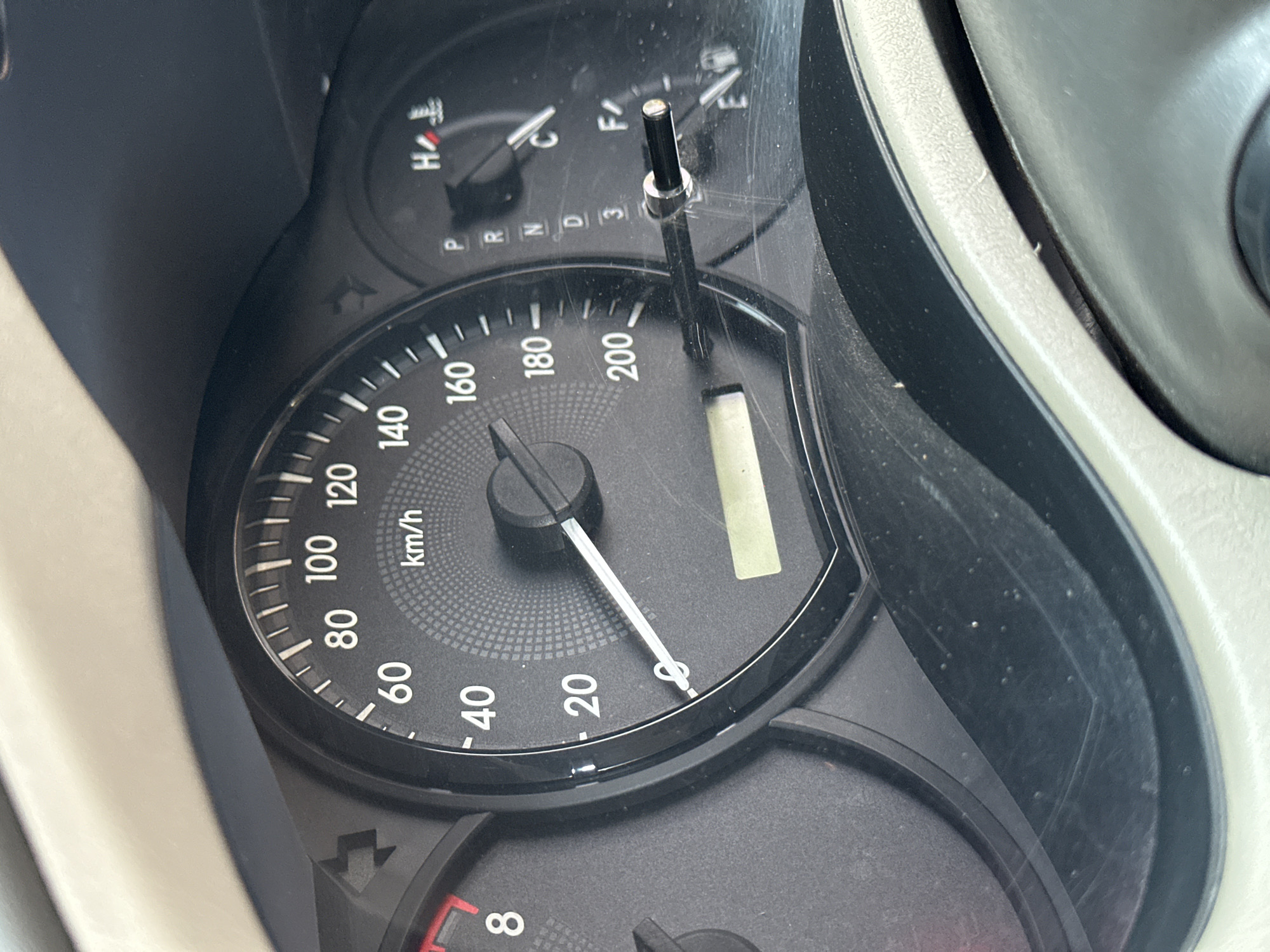 The image shows a close-up view of a car's dashboard, specifically the speedometer and other gauges. The speedometer displays a speed of approximately 20 km/h. To the right of the speedometer are other indicators, including a temperature gauge showing the engine is slightly above normal operating temperature (indicated by the red mark on 'H'), a gear selector showing the car is in drive ('D'), and a fuel gauge showing the tank is close to empty ('E'). The dashboard is made of dark plastic and shows some signs of wear, including scratches and dust. The background is out of focus but appears to be the interior of a car. The image was taken during the daytime, likely by the driver, as evidenced by the angle and framing. The location in the city of Bulakrejo, Indonesia, is not explicitly visible in the image, but the context of a car dashboard suggests it was taken within the urban environment of that city. There are no people visible in the image.