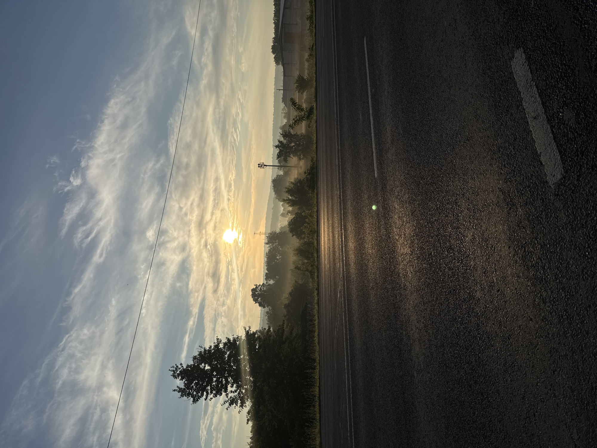 This image depicts a tranquil rural roadside scene at sunrise, likely located near Strășeni, Moldova. The sun is low on the horizon, casting a warm, golden light across the landscape. Wispy clouds streak across a clear blue sky, illuminated by the rising sun. A single power line stretches horizontally across the upper portion of the frame.
In the foreground, a dark asphalt road occupies the lower half of the image, with a white dashed line visible on the right side. The road surface appears wet or reflective, catching the sunlight. To the left of the road, a dense line of trees forms a dark silhouette against the brighter sky. A large, leafy tree stands prominently on the left, its branches silhouetted.
Beyond the road and the trees, a gentle mist or fog hangs low over the fields and hills, partially obscuring the distant landscape. The sunlight filters through the mist, creating a soft, diffused glow. Several utility poles and possibly some agricultural structures or buildings are visible in the mid-ground and background, hinting at the proximity of a rural settlement. The overall impression is one of quietude and the beginning of a new day in a serene, natural environment. There are no people or animals visible in the image.