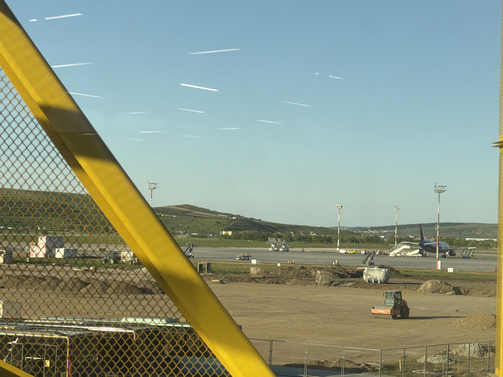 The image shows a construction site at Cluj International Airport, Cluj-Napoca, Romania, taken through a yellow metal mesh structure. In the background, an airplane is parked on the tarmac near terminal equipment. In the mid-ground, there is a construction site with piles of dirt and heavy equipment. There is a hill with green grass visible in the background. The sky is light blue with several airplane contrails.