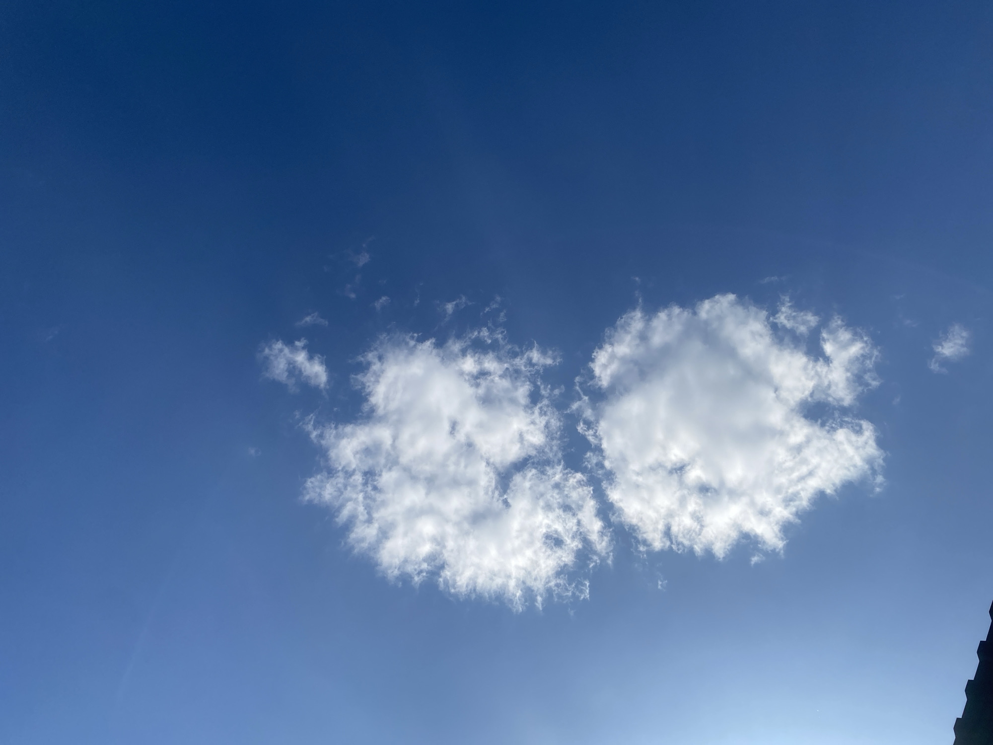 This image shows a clear blue sky with two fluffy white clouds. In the bottom left corner, a dark, angled object, likely the edge of a roof, is visible. The clouds are scattered and appear to be cumulus clouds. The overall environment suggests an outdoor scene under a sunny sky in a location that could be rural or urban. There are no people or animals present in the image. The scene is static and appears to have been captured on a bright day.