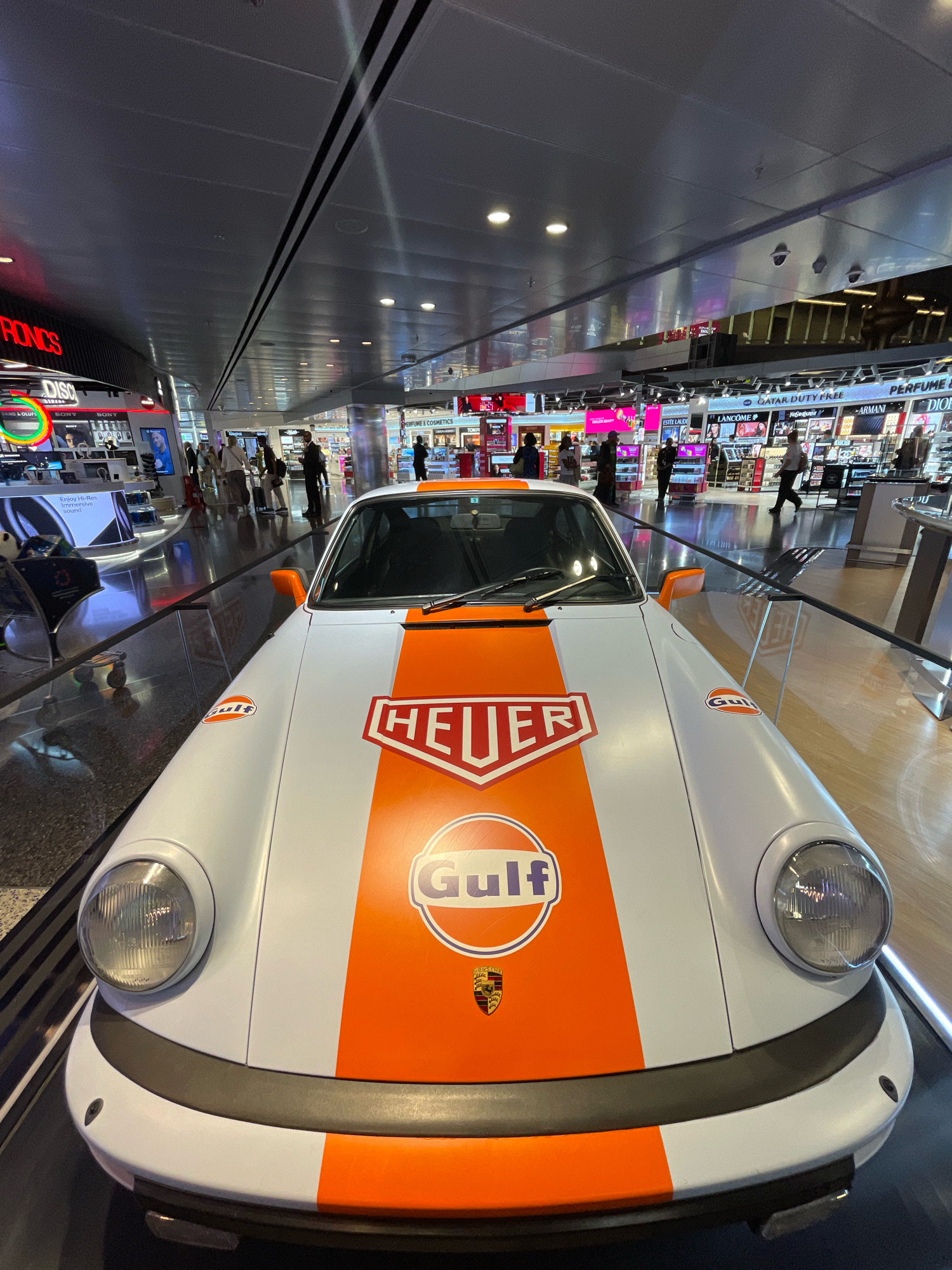 The image shows a white and orange Porsche 911 race car displayed in a duty-free shop, likely at Hamad International Airport in Doha, Qatar. The car features prominent "Gulf" and "Heuer" branding. In the background, shelves stocked with perfumes and cosmetics are visible, along with various shoppers and airport staff. The polished floor reflects the bright lighting and displays.