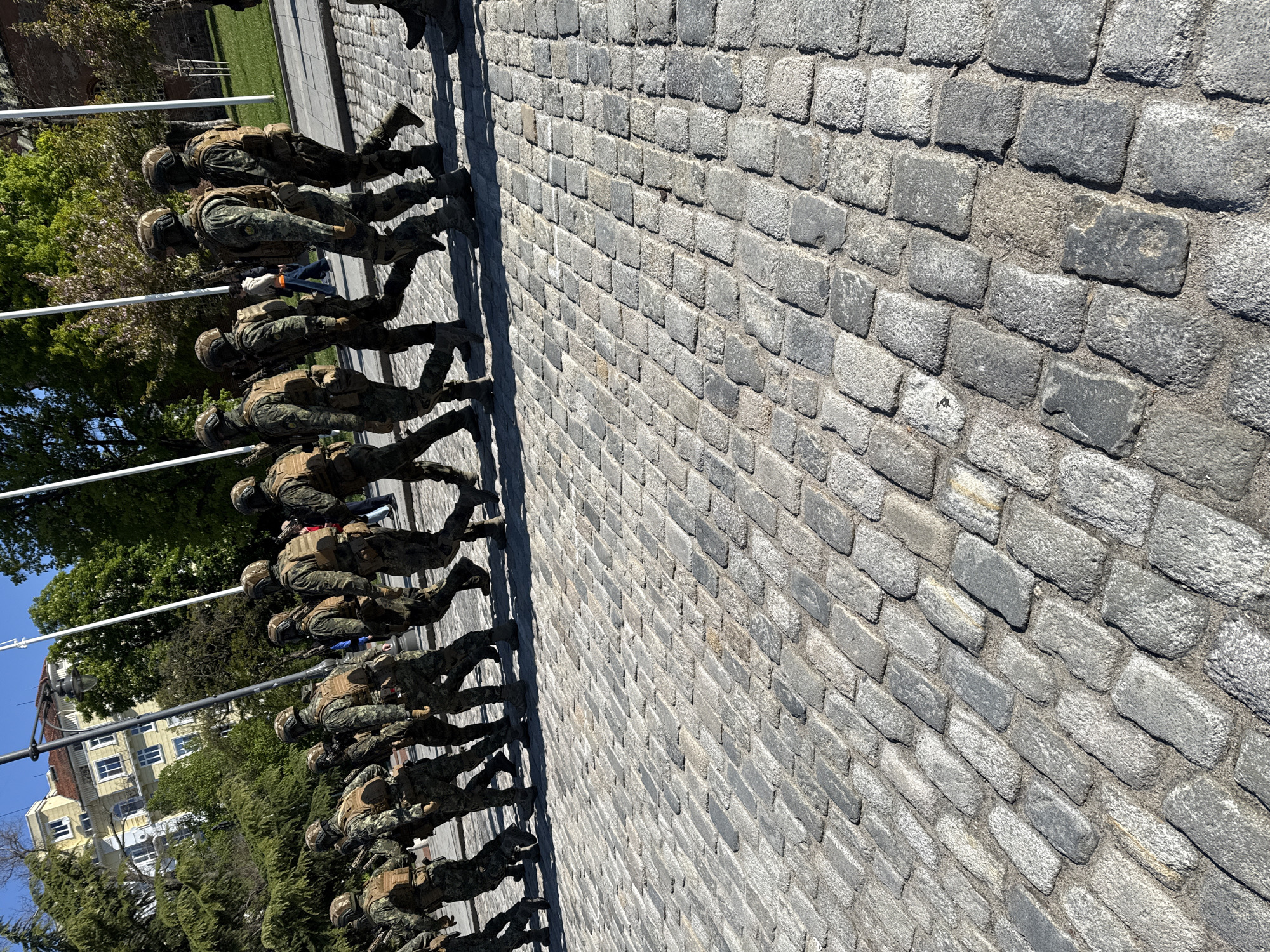 The image shows a group of soldiers marching in formation on a cobblestone street. The location is likely in Sofia, Bulgaria, based on the architectural style of the buildings and the vegetation in the background. The soldiers are wearing camouflage uniforms and helmets, and they are carrying rifles. They are marching in a straight line along the edge of the cobblestone path. In the background are trees, a building with several visible windows, and traffic lights. The lighting suggests it is daytime with bright sunlight.