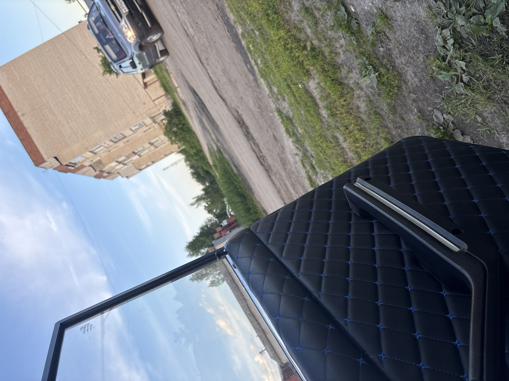 The image captures a close-up view of a car's interior door panel, featuring black quilted upholstery with blue stitching, and a chrome-trimmed handle. The open car door reveals a glimpse of the exterior, with a clear blue sky dotted with wispy clouds. In the background, a multi-story brick building stands, with a dark-colored SUV parked on a cracked asphalt road. Patches of green grass and weeds are visible along the roadside. The scene appears to be set in an urban or suburban environment, possibly during daylight hours.