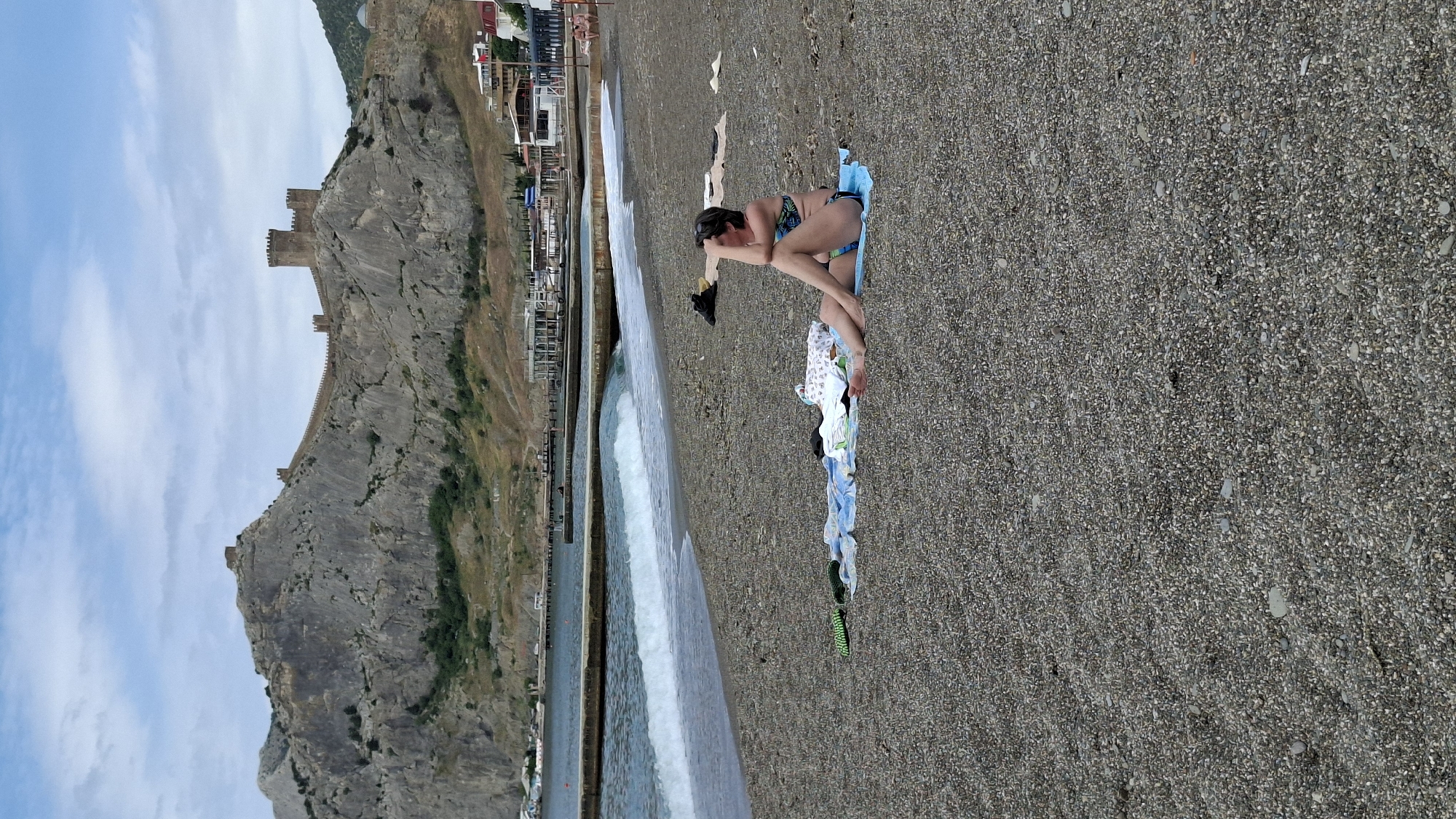 A daytime scene captures a pebble beach alongside a body of water with a large rocky cliff or mountain in the background. The sky is partly cloudy with patches of blue.
On the pebble beach, a woman in a two-piece swimsuit (bikini) is lying on a blue towel. She is positioned with her torso slightly elevated, resting on her arms, and her legs bent. Next to her, several items of clothing or towels are spread out on the pebbles. Black footwear is also visible near her. The beach surface is composed of small, dark gray and light-colored pebbles.
To the left of the beach, a body of water, likely the sea, extends. Waves are breaking gently near the shore, creating a white foam line. Further out, the water appears calm. Along the far side of the water, distant buildings and structures are visible on what seems to be another part of the coastline or a harbor area.
Dominating the background is a massive, craggy rock formation or mountain. Perched atop this rocky landscape are the visible walls and towers of an ancient fortress or castle, made of stone. The fortress structure runs along the ridge of the mountain. The slopes of the mountain show some sparse vegetation.
The overall impression is one of a coastal resort or historical site, with natural beauty and historical elements coexisting. There is no visible text in the image.