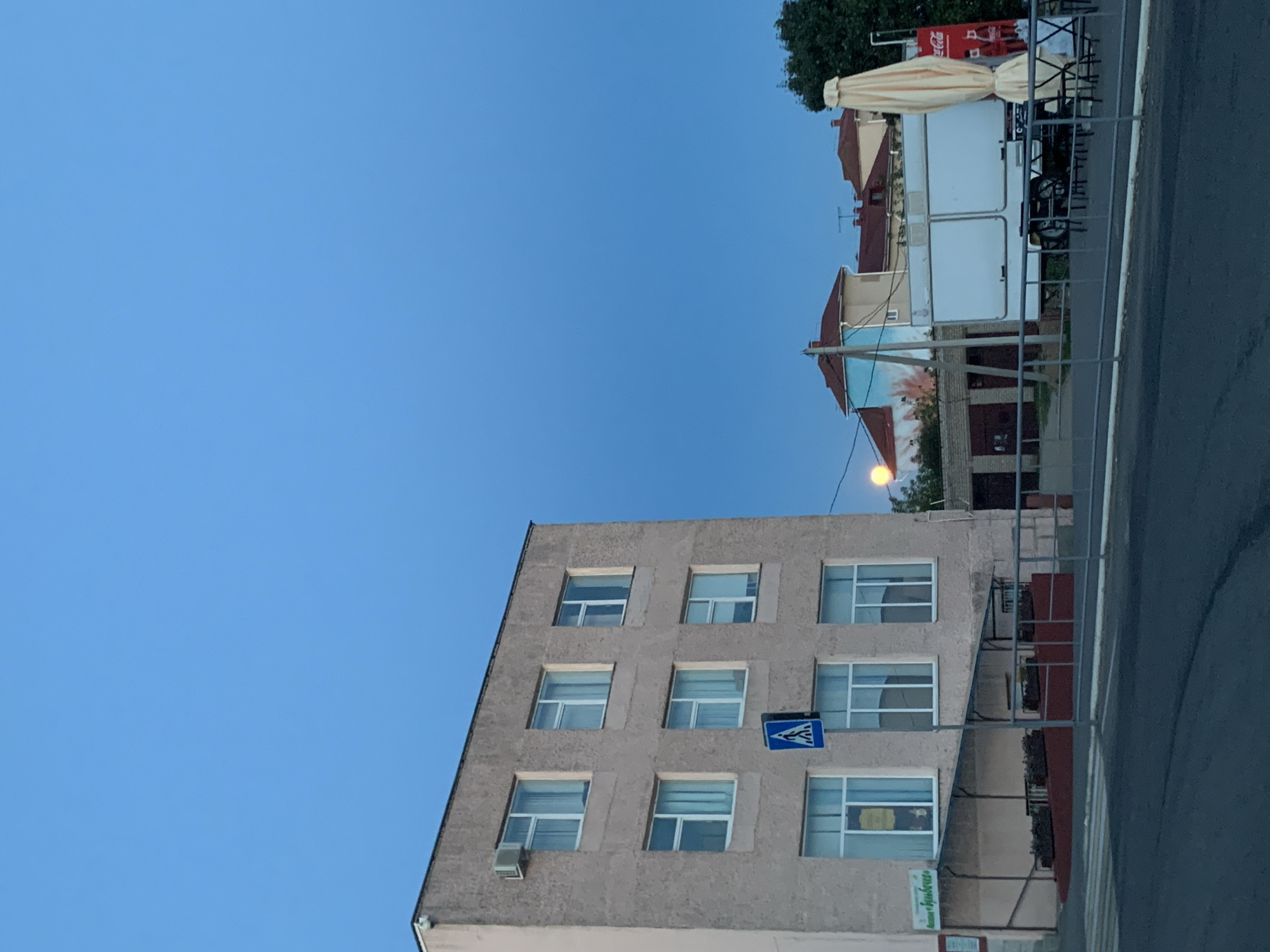 The image depicts a street scene in Kapyĺ, Belarus, under a clear, deep blue sky. In the foreground, a paved road is visible, with a pedestrian crossing marked by white stripes. A silver metal railing separates the road from the sidewalk area.
Dominating the left side of the image is a three-story building with a light-colored, textured facade. The building has multiple windows, some of which are open with curtains visible. An air conditioning unit is mounted on the upper left side of the building. A sign with green lettering is affixed to the lower left facade. A blue pedestrian crossing sign is mounted on a pole in front of the building. A ramp leads up to the building's entrance, bordered by planters with red mulch.
To the right of the building, further back, is a white trailer-like structure with two large windows or doors. Behind this structure, a tall, closed umbrella, a few outdoor tables and chairs, and a Coca-Cola advertisement are visible. The background includes parts of other buildings with reddish-brown tiled roofs and trees. A bright, orange, circular light source, possibly a street lamp or the setting sun, is visible on the far right of the frame. The overall impression is a quiet urban environment during twilight.