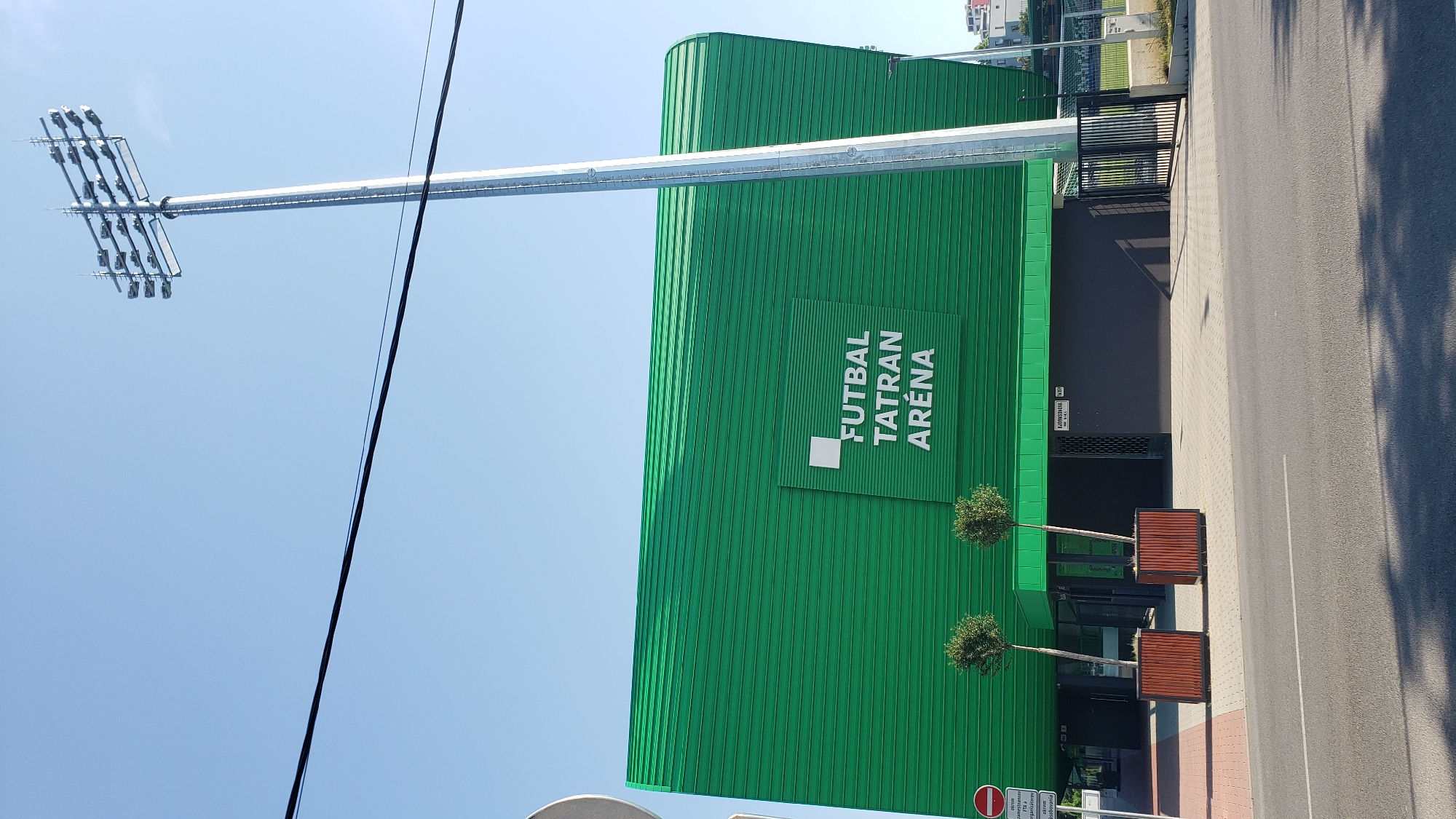 The image depicts the Futbal Tatran Aréna stadium in Prešov, Slovakia. The stadium features a distinctive bright green, corrugated metal facade. In front of the building, there are two planter boxes with small, spherical trees. A tall, silver lighting tower is prominently visible to the right of the stadium. A black metal gate is also present, leading to an outdoor area that appears to be a sports field. The scene is set on a clear, sunny day with a blue sky and some overhead power lines. The foreground shows a paved sidewalk and a street, with shadows cast by the stadium and trees. A street sign with Slovakian text is visible near the entrance.