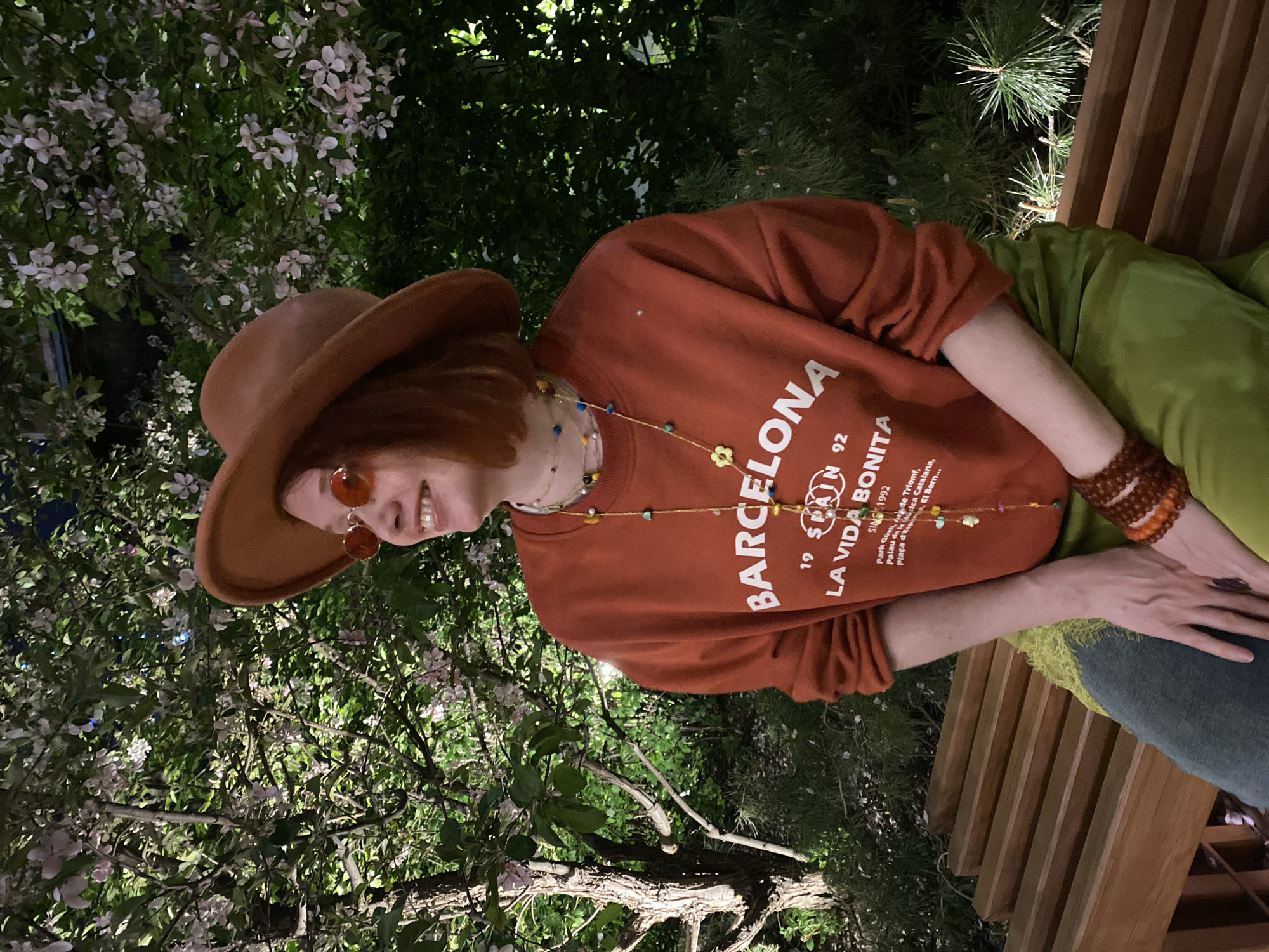 A person wearing a brown hat, sunglasses, and an orange sweatshirt with "BARCELONA 19 SPAIN 92 LA VIDA BONITA SINCE 1992" printed on it sits on a wooden bench. The person also wears a colorful necklace and beaded bracelets. Behind the person are flowering trees and green bushes. The scene does not appear to be in Yekaterinburg, Russia, based on the clothing, text on the sweatshirt, and the flowering plants.
