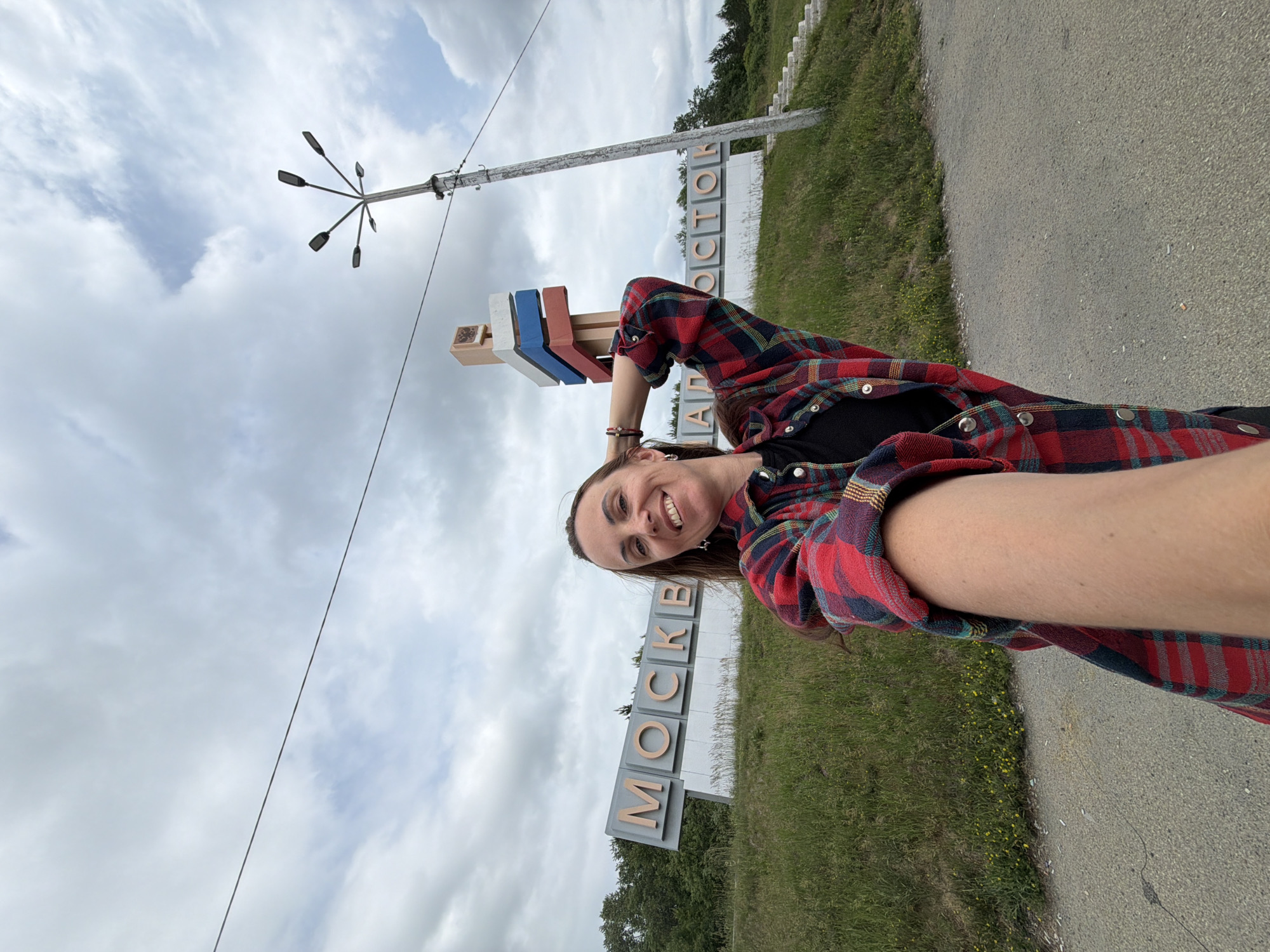A woman stands on a paved road, taking a selfie. She is wearing a red and blue plaid shirt over a black t-shirt and has her left arm raised, with her hand behind her head. She is smiling and looking at the camera.
Behind her, a large sign with the word "MOSCOW" in large, metallic letters is visible. To the right of the sign, a lamppost with multiple lamps extends upwards, and a smaller sign with Russian flag colors is attached to a pole. The environment is grassy with some yellow wildflowers. The sky is cloudy, with patches of blue visible. The road surface is asphalt.
