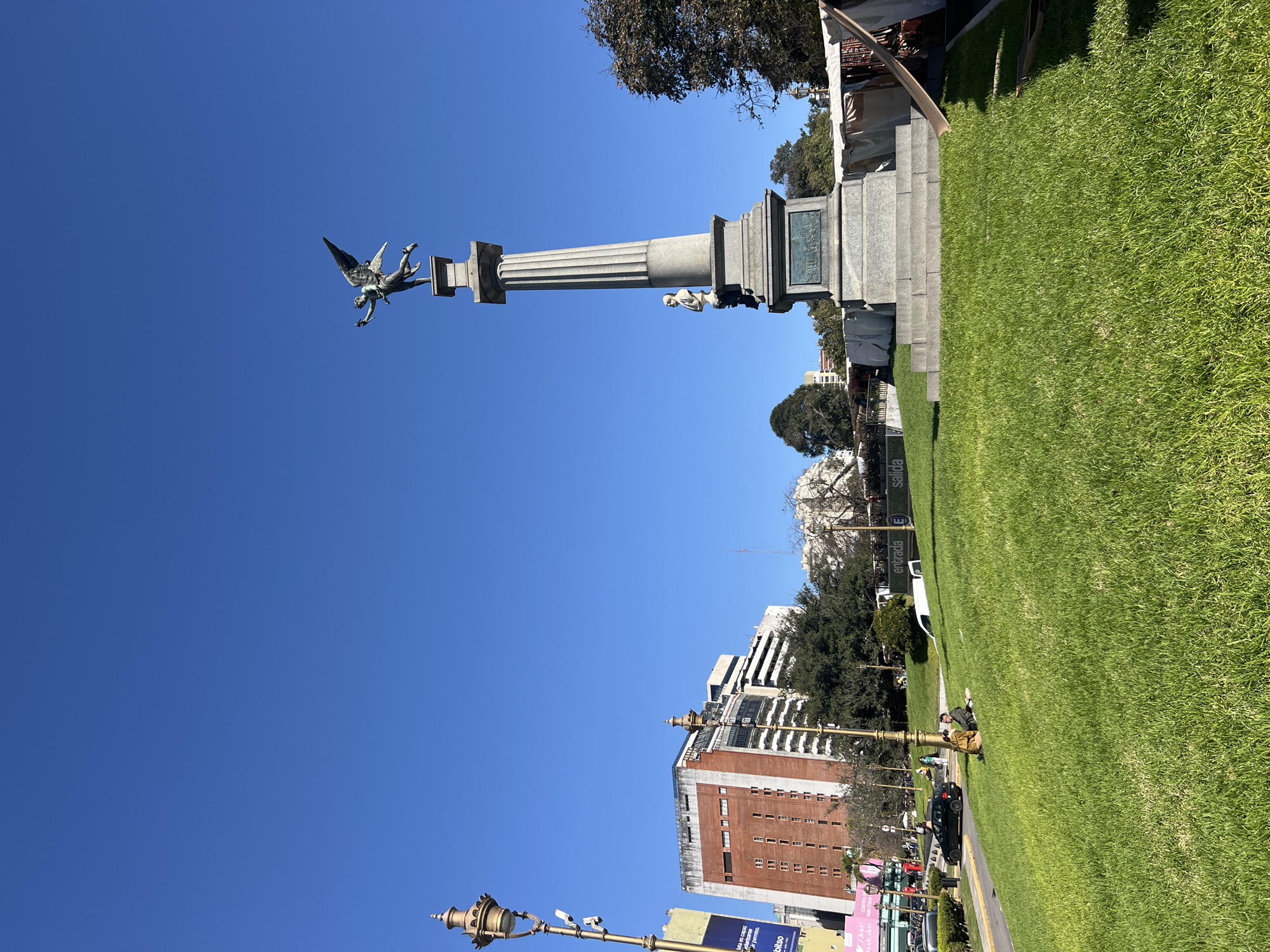The image captures a bright, sunny day in Buenos Aires, Argentina, featuring the Columna del Centenario, a monument topped with a winged figure, likely Icarus. The monument stands on a grassy area in Plaza Alemania. Buildings are visible in the background, indicating an urban setting. Below the monument, a sidewalk runs parallel to the grass, where people are relaxing. Light poles are seen in the foreground. The scene is bathed in sunlight, creating sharp shadows.