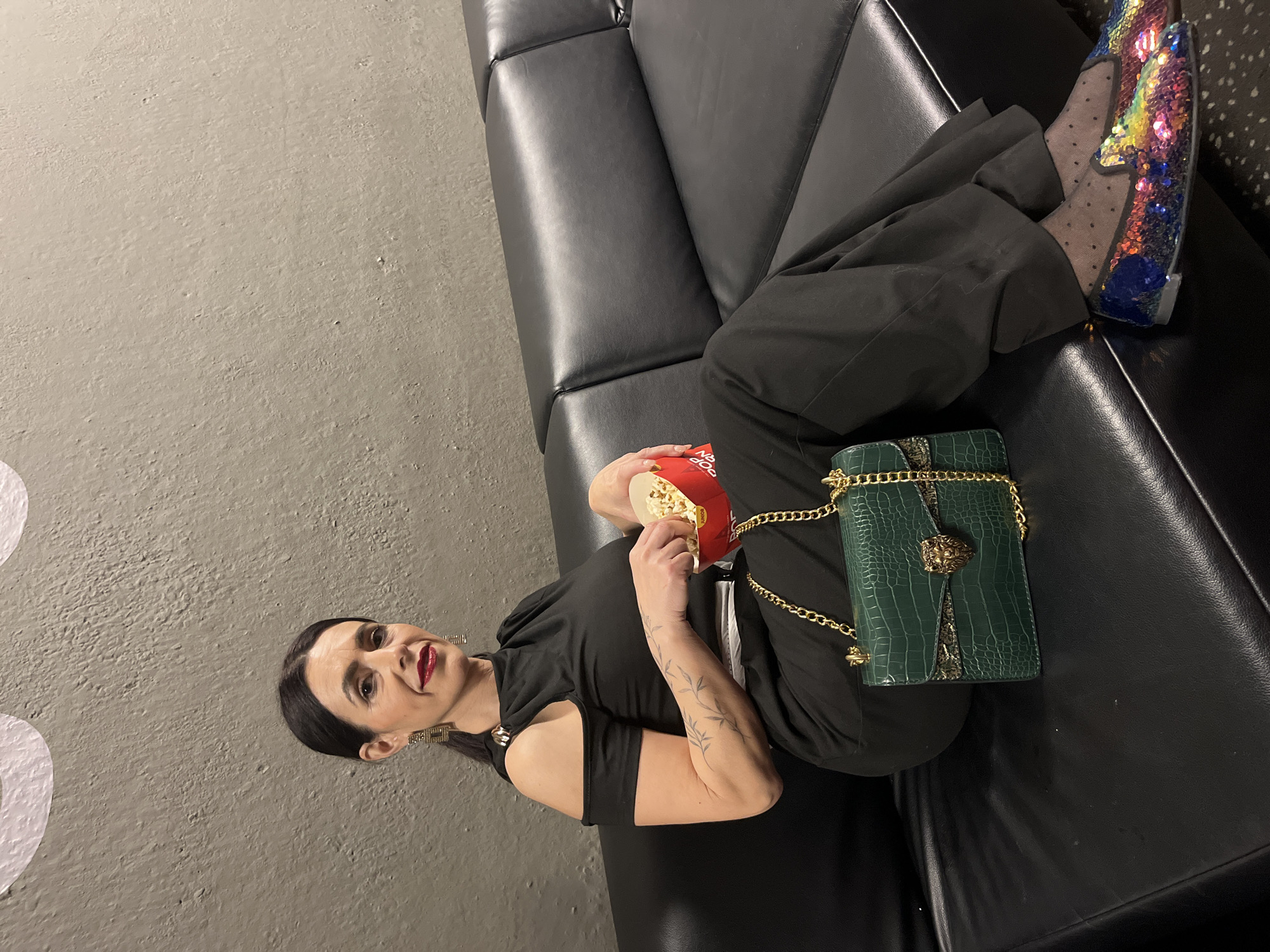 A woman is reclining on a black leather sofa, holding a red container of popcorn in her hands. She is wearing black trousers, a black top with a cut-out shoulder, and sequined shoes. She also has a green crocodile-patterned purse with a gold chain. Her dark hair is pulled back, and she is wearing large square earrings and red lipstick. A tattoo of leaves is visible on her left arm. The sofa is in front of a gray wall. The setting could be a cinema in the city of Luxembourg, Luxembourg.