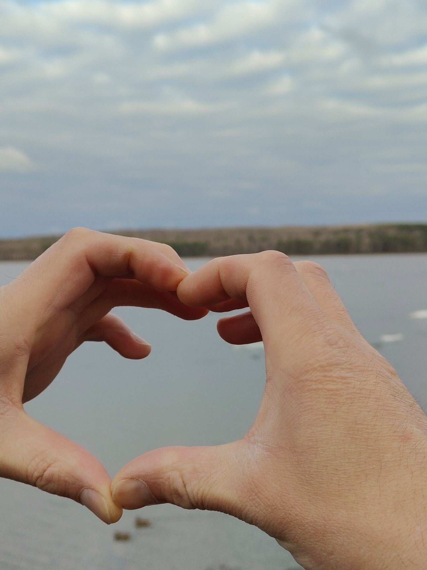 The image depicts a pair of Caucasian hands forming a heart shape against a backdrop of a body of water, possibly a lake or a wide river. Beyond the water is a line of trees. The sky is overcast with grey clouds. The location is Unknown, Unknown. No specific individuals or actions are discernible other than the hands forming a heart shape.