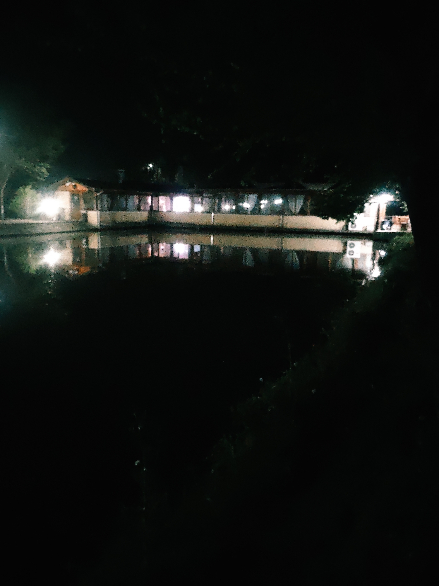 The media file presents a nighttime scene centered on a long, low-rise building illuminated by artificial lights, situated adjacent to a dark body of water. The building, possibly a restaurant or event venue, features multiple windows or open sections, many of which display white curtains or blinds. A smaller, separate structure is visible on the far left, also brightly lit.
The primary activity suggested is the operation of the establishment, indicated by its illumination. The bright lights from the building cast reflections onto the surface of the water, which appear slightly distorted, suggesting gentle ripples. The surrounding environment is characterized by dark vegetation, likely trees or bushes, framing the scene on both sides and receding into the blackness of the night sky. The time of day is clearly night, with no natural light visible. The atmosphere appears calm and serene. No people or specific events are discernible. There is no visible text in the image.