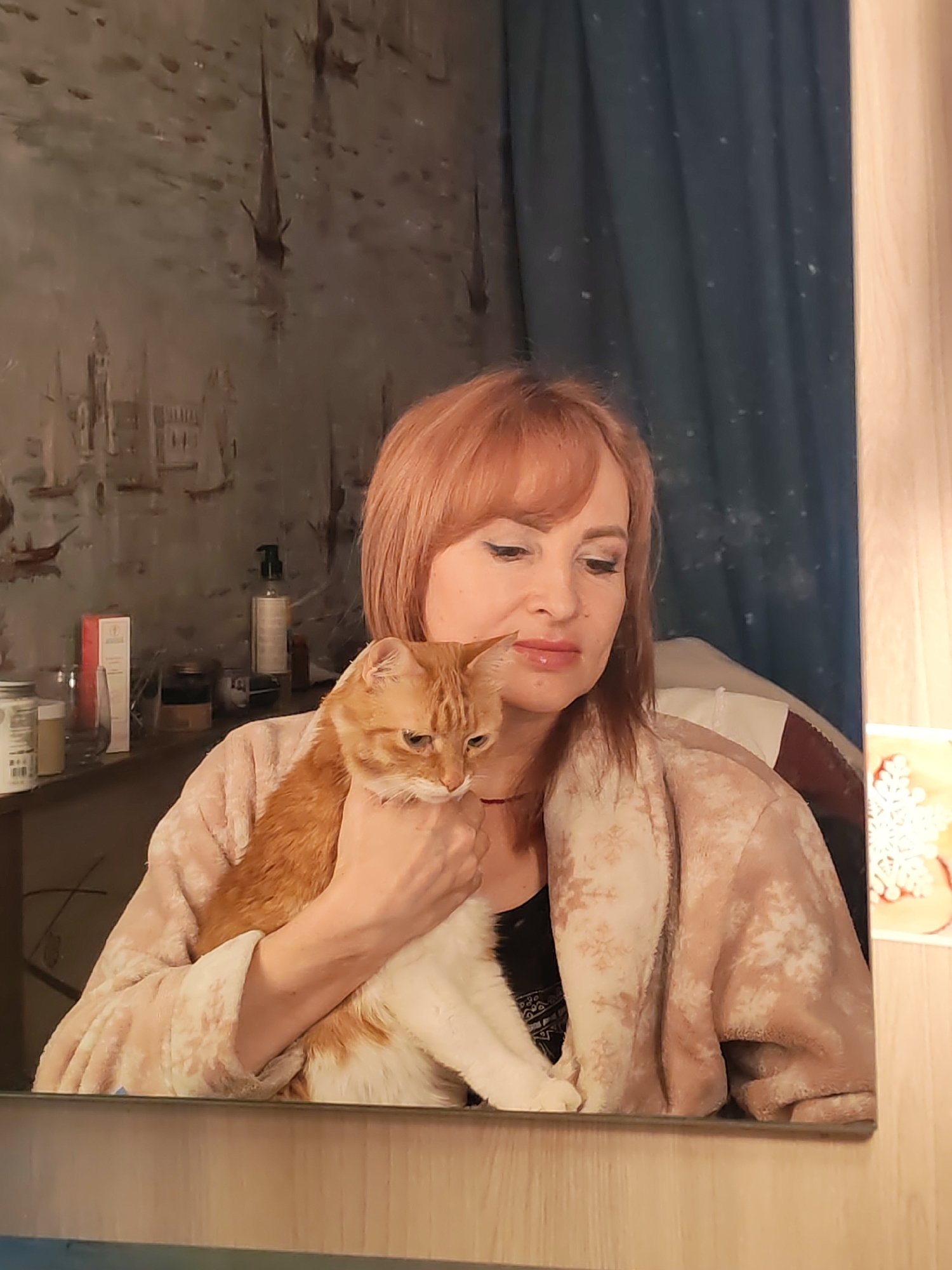 A red-haired woman in a light beige robe with snowflake patterns is holding a ginger and white cat. The woman has bangs and is wearing makeup. Behind her is a wall with a cityscape painting, a shelf with various bottles and containers, and a blue curtain. The image appears to be a reflection in a mirror. The location is possibly Chaikovsky, Russia.