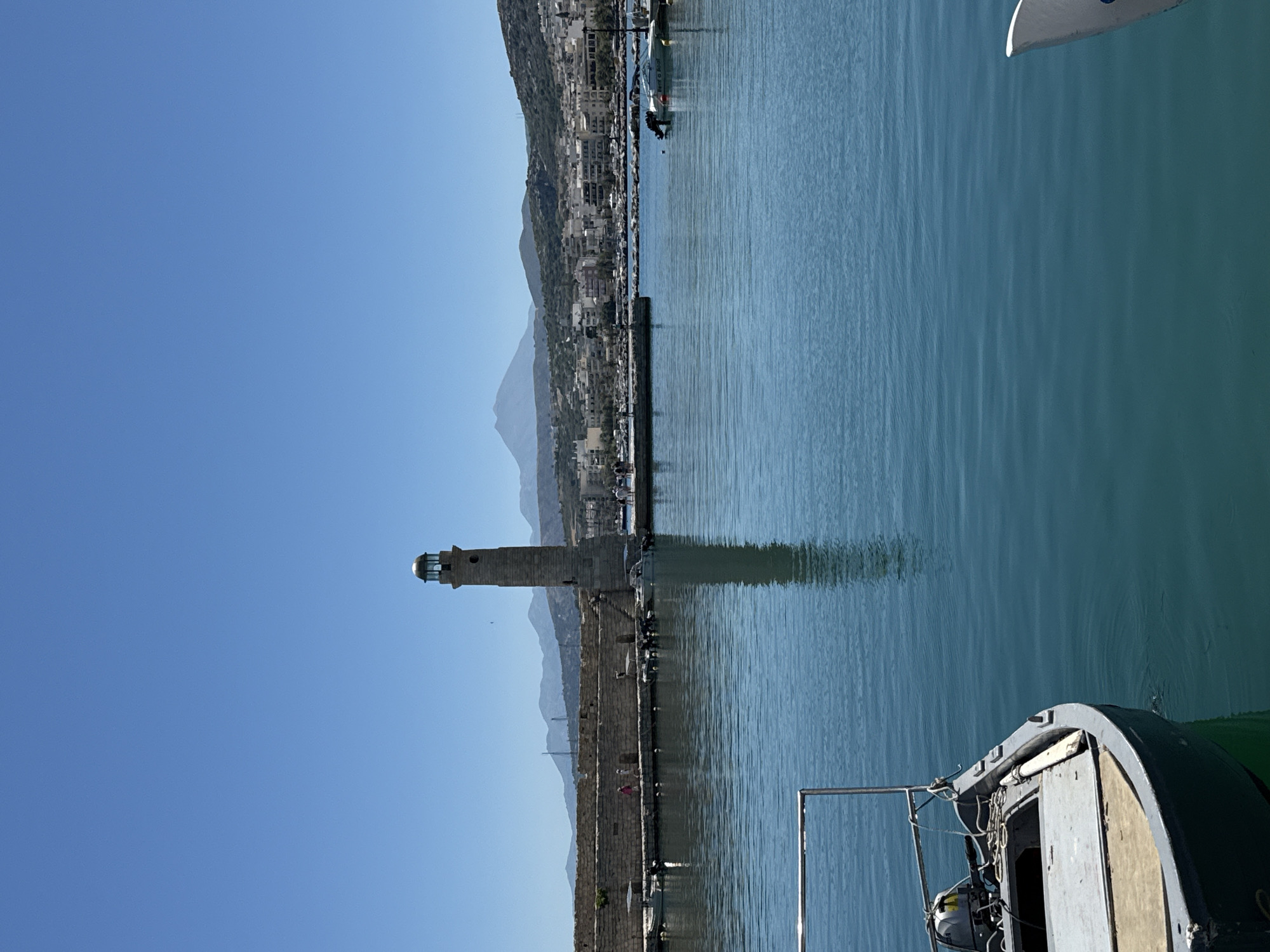The image depicts the Venetian harbor of Rethymno, Crete, Greece, on a clear, sunny day. In the foreground, a small, gray motorboat is moored, its bow extending into the lower-left quadrant of the frame. The boat has a weathered appearance with visible wear and tear. The turquoise water of the harbor is calm, with gentle ripples reflecting the bright blue sky.
Dominating the mid-ground is the iconic Venetian lighthouse, a tall, stone structure with a distinctive lantern room at its apex, situated at the end of a long, stone quay. The quay is lined with people strolling and several small boats are docked along its edge. The stone fortifications of the harbor wall are visible, with a weathered, ancient appearance.
In the background, the city of Rethymno unfolds, a dense collection of buildings with white and earth-toned facades. Behind the city, a range of mountains rises, with one prominent peak appearing somewhat conical and distant. The overall scene conveys a sense of a peaceful, sun-drenched coastal town.