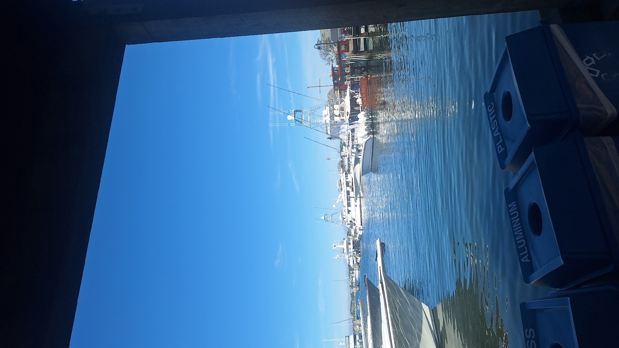 The image captures a view of the Nantucket harbor on a clear day, with several boats docked in the blue water. In the foreground, three blue recycling bins are visible, labeled "GLASS," "ALUMINUM," and "PLASTIC." The bins are positioned near the water's edge, suggesting a public area or boardwalk. The middle ground is dominated by various boats, including large yachts and smaller fishing vessels, all appearing to be moored. The background shows a more distant shoreline with buildings and trees, indicative of the town of Nantucket. The sky is a vibrant blue with a few wispy clouds. The overall scene depicts a peaceful and sunny day at a well-maintained harbor.