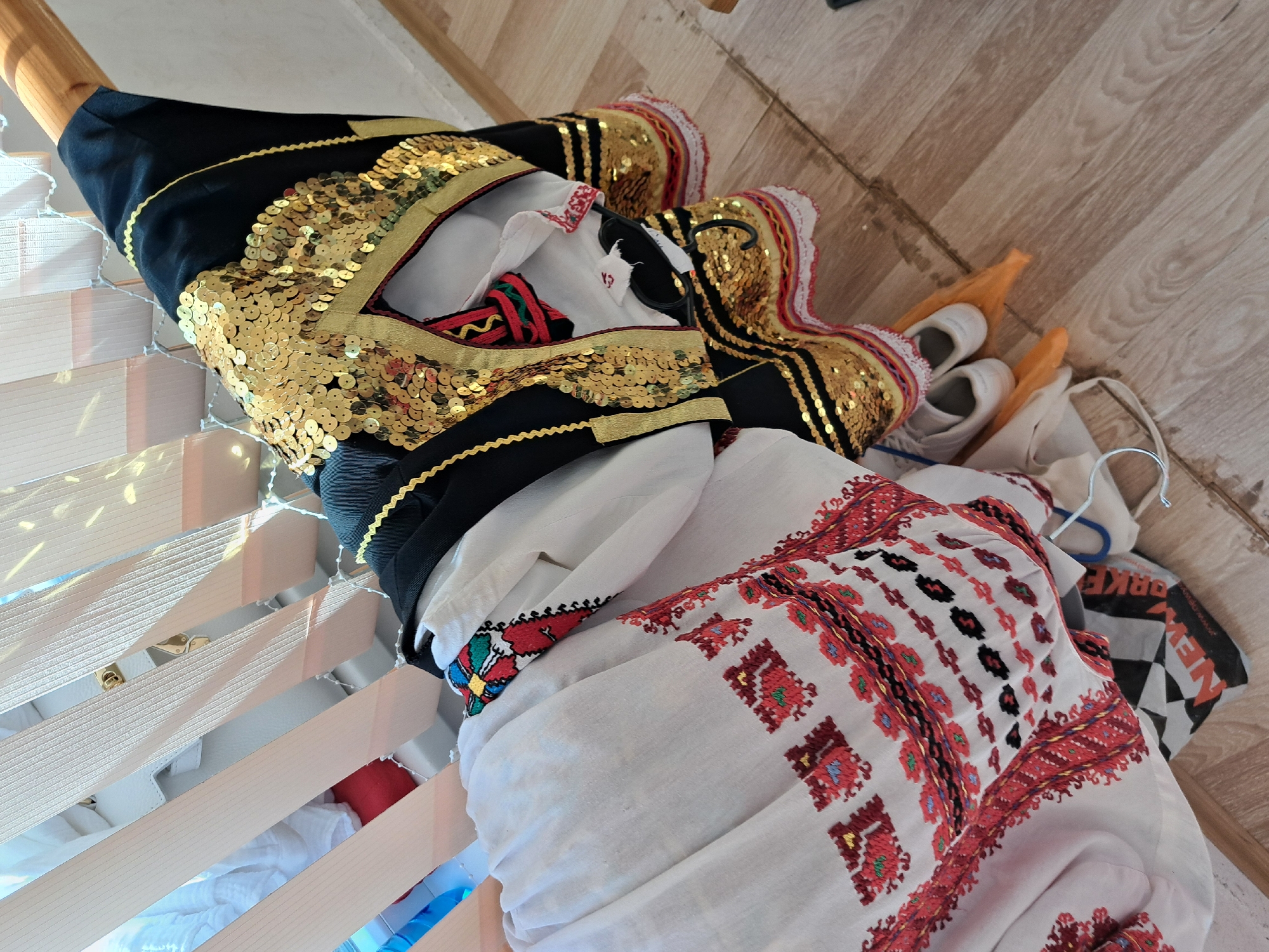 The image shows a traditional Bulgarian folk costume laid out on what appears to be a bed or a storage unit with wooden slats. The costume includes a white embroidered shirt with red and black patterns, and a black vest or bodice with gold sequin embellishments. There are also small white shoes and other items, like a bag with a checkered pattern, nearby. The location is likely within a domestic setting, but there is no information in the image to confirm that it is in Lokorsko, Bulgaria.