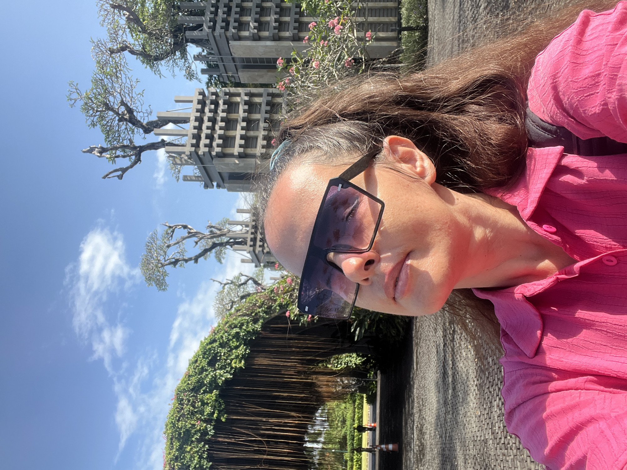 In Kuta Selatan, Indonesia, a woman takes a selfie outdoors. She is looking up at the sun, wearing large sunglasses, a bright pink collared shirt, and has her brown hair pulled back in a ponytail. Behind her, a large archway covered in dense green vines with hanging roots is visible. Further in the background, there are unusual concrete structures with stacked layers resembling a castle, topped with trees. The ground is paved with cobblestones, and the sky is blue with scattered white clouds.