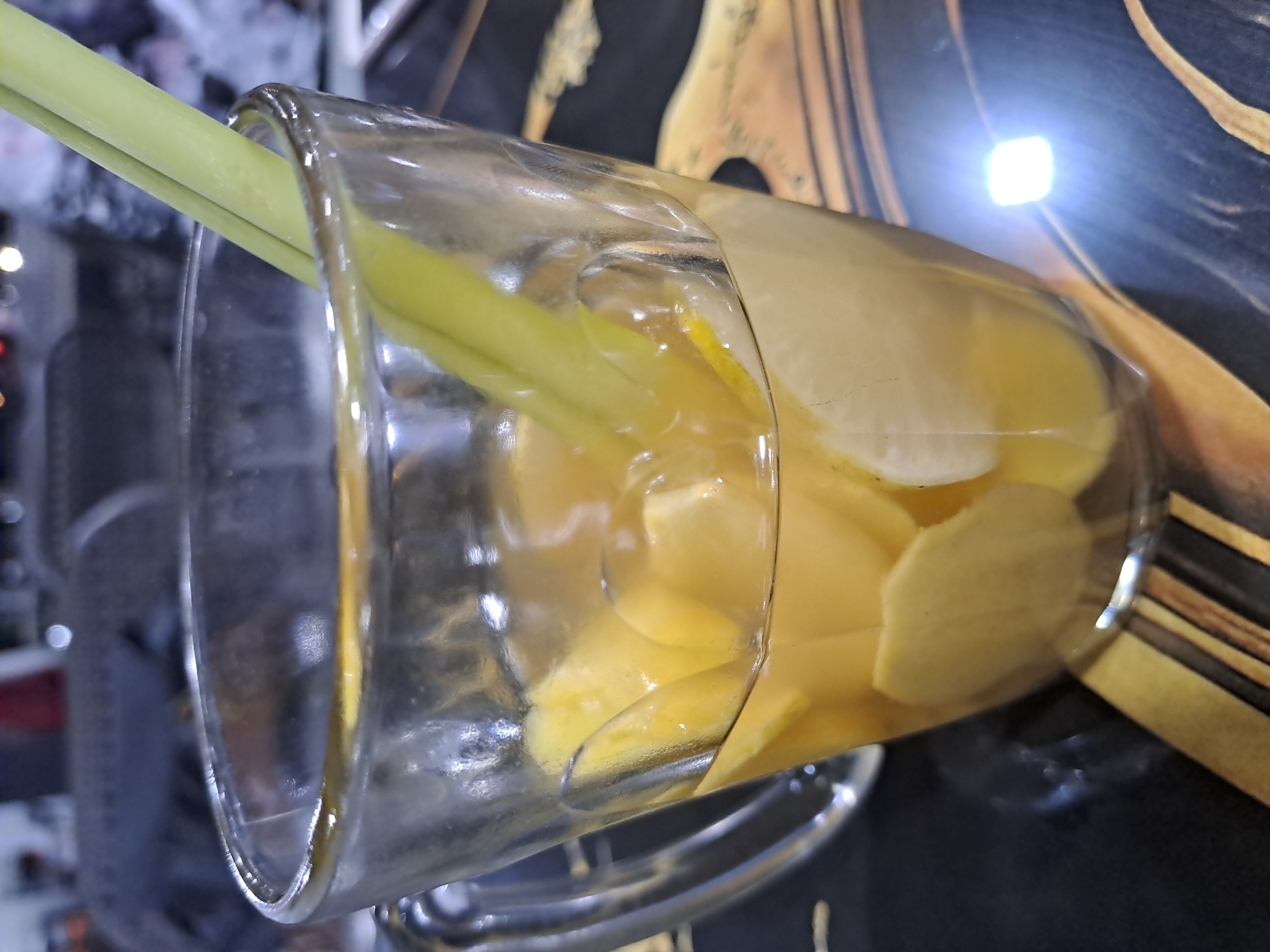A clear glass pitcher or mug, rotated 90 degrees clockwise, is filled with a pale yellow liquid and various ingredients. Submerged in the liquid are numerous thin, light yellow slices, strongly resembling ginger. Two long, light green stalks, consistent with lemongrass, protrude from the top opening of the pitcher. A thin, curved slice of what appears to be lemon or lime is also visible near the upper right surface of the liquid.
The background is dark and largely out of focus, suggesting an indoor environment. A bright, distinct square-shaped light source creates a strong reflection in the upper right background, casting a glow on the glass and surrounding area. The glass itself shows reflections of the blurred surroundings. The image captures a beverage, likely an infused drink or herbal tea, with ingredients commonly used in Indonesian cuisine. There are no people visible, nor is there any discernible text. The focus is on the contents of the pitcher and the play of light on the glass.