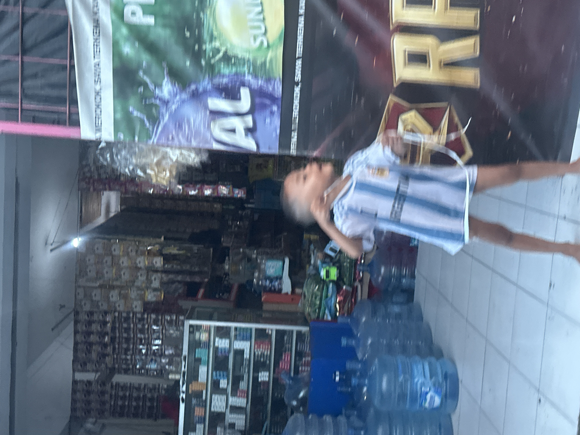 A young boy, wearing an Argentinian soccer jersey, stands barefoot in front of a store in Kuta, Indonesia. The store appears to be a small convenience store or warung. Behind the boy, there are shelves stocked with various items, including what appear to be cigarette packs and bottled drinks. A large banner with text in Indonesian, possibly advertising a product or event, is visible to the right of the boy. The floor is tiled and a row of large water jugs is lined up to the left of the scene. The image is slightly blurred due to motion.