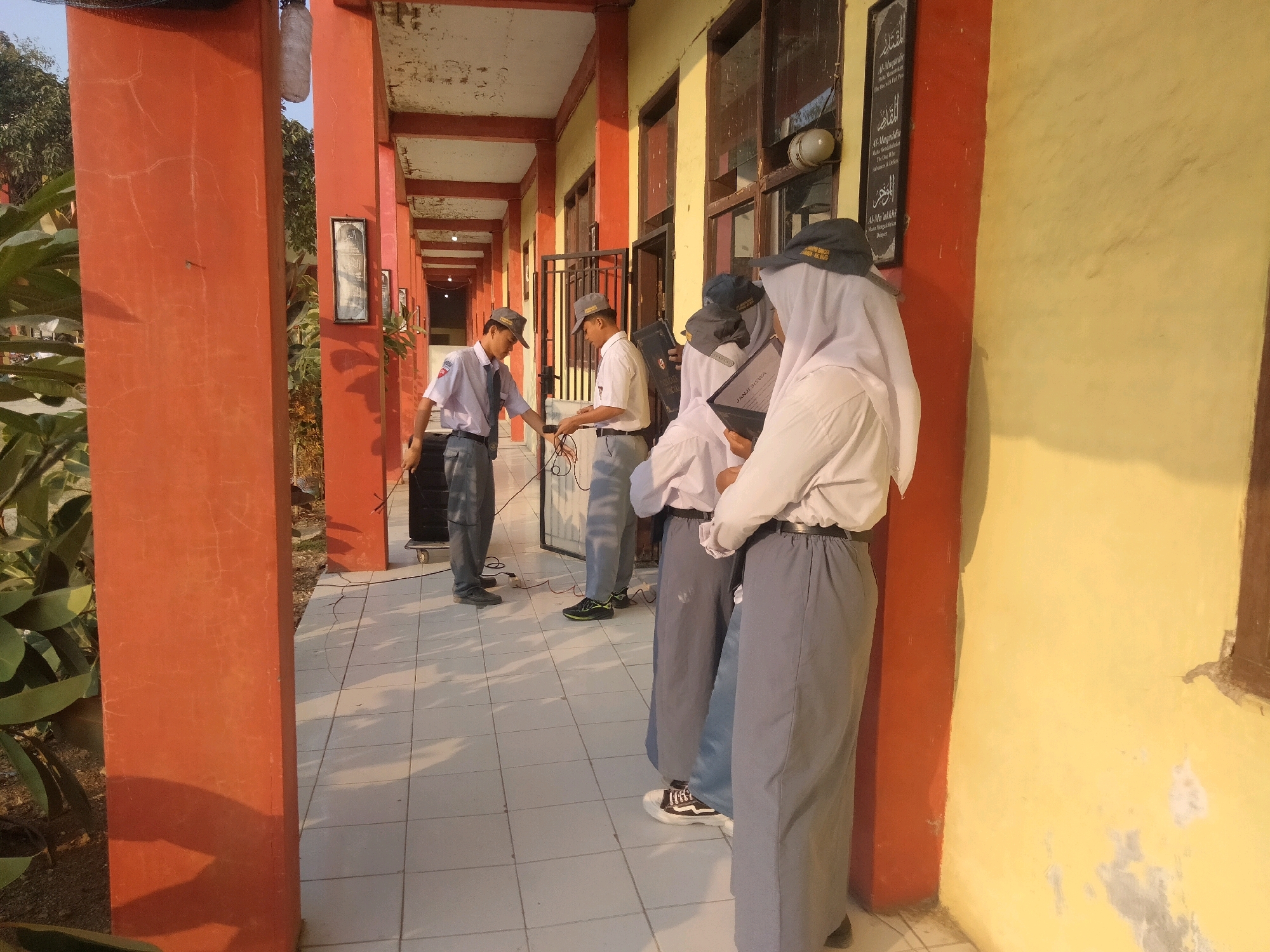 The image depicts a school hallway in Suka Sari, Indonesia, with students in uniform. On the left, a male student in a white shirt, tie, and grey pants stands near a speaker, holding a cable. To his right, another male student in a white shirt and grey pants is adjusting something on a table. Further down the hallway, three students, two with grey hats and one wearing a white hijab, are gathered, with two of them holding books. The hallway has orange pillars and a yellow wall with windows. The ground is tiled. The scene appears to be outdoors during daylight.