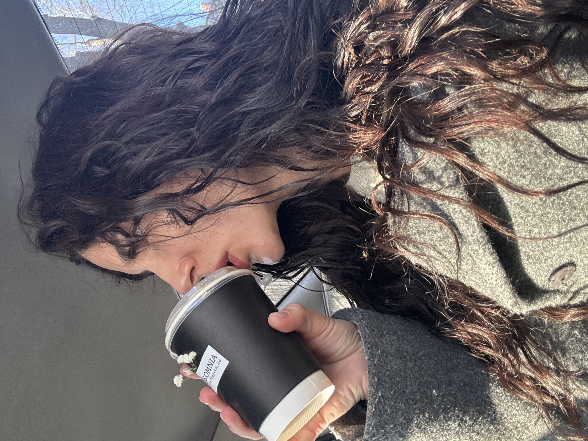 An individual with long, dark, wavy hair is depicted in profile from the right, drinking from a black paper cup with a clear plastic lid while seated inside what appears to be a vehicle. The cup, held by the person's left hand, features a white label reading "SOMNIA" and "на Маркса, 26". Small white decorative flowers are tucked under the cup's lid on the left side. The person is wearing a gray, textured coat. In the upper right background, through a vehicle window, bare tree branches are visible against a bright blue sky. The address "на Маркса, 26" indicates a location on Marx Street in the city of Chaikovsky, Russia.