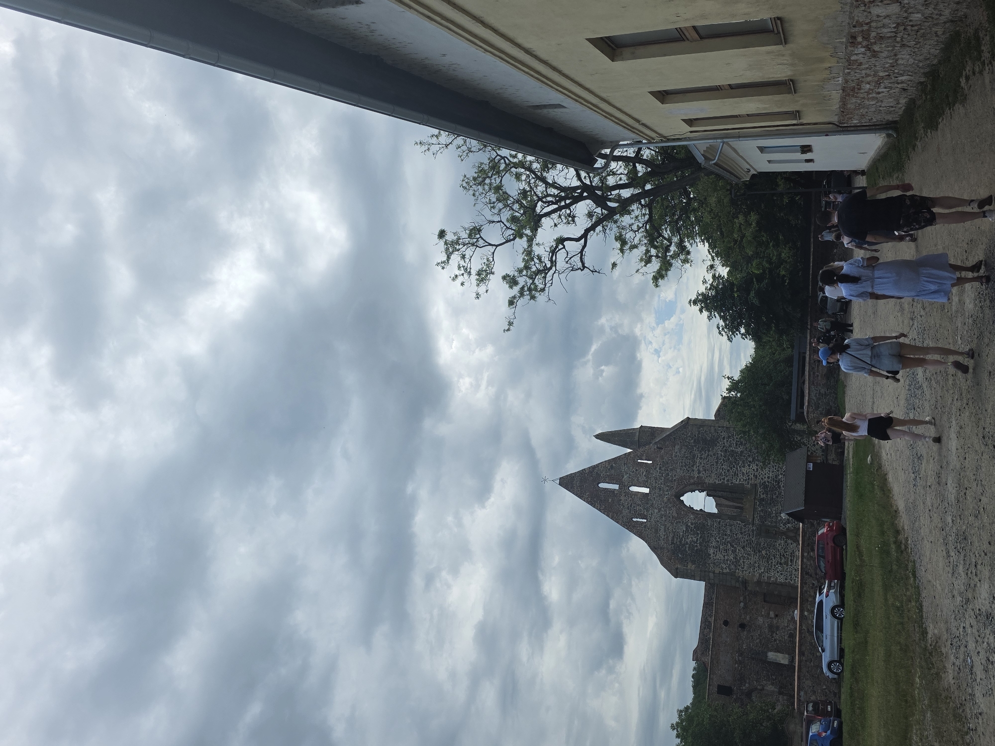 The image depicts a partially obscured view of a stone ruin against a cloudy sky. The ruin has a pointed archway. To the right of the ruin is a dirt path with people walking on it. Several cars are parked nearby. The top of the image includes a building overhang with a tree branch extending towards the center of the scene. The location of the image is Unknown, Unknown.
