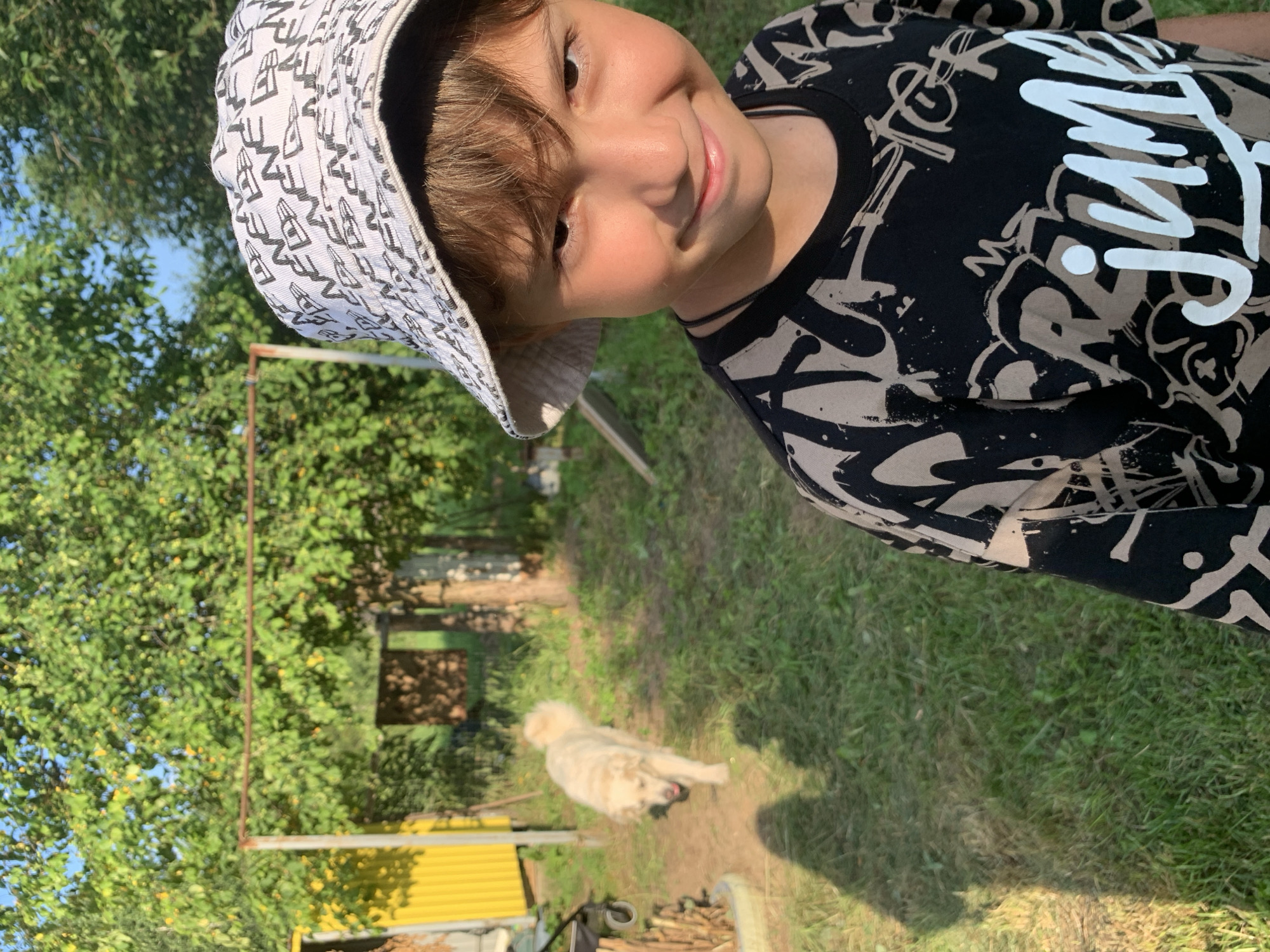 In Zasechnyi, Russia, a young boy wearing a bucket hat and a black t-shirt with a graphic print stands in a grassy outdoor area. Behind him, a light-colored dog walks across a dirt path. The background features trees with green foliage and some yellow siding of a structure. Sunlight casts shadows on the ground.