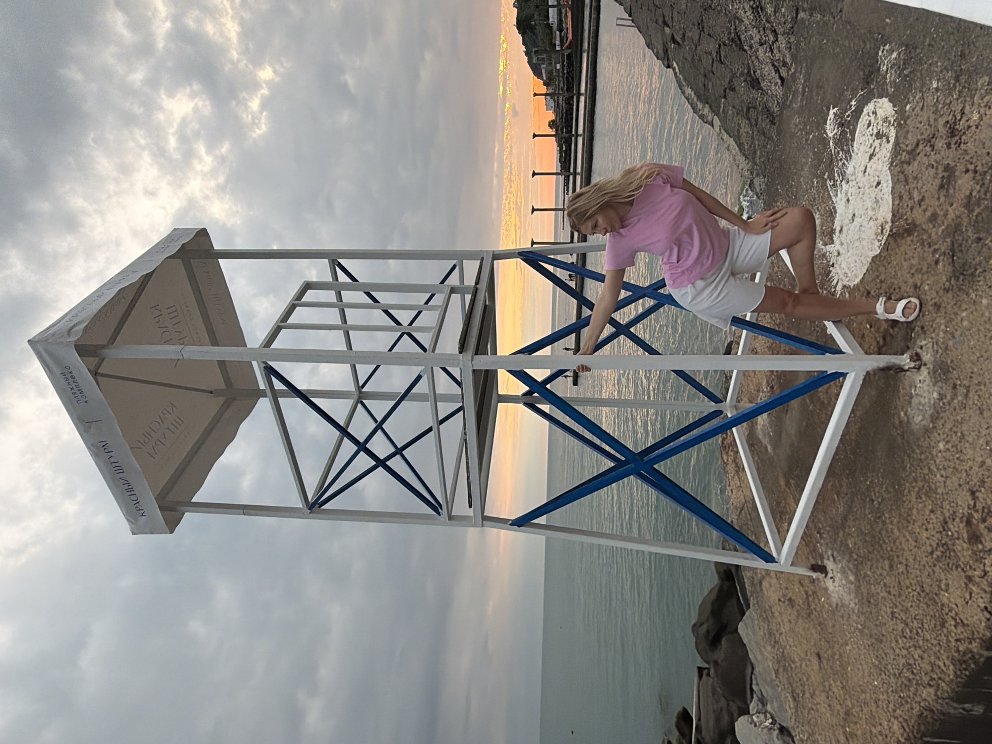 A young woman with blonde hair, wearing a pink t-shirt and white shorts, poses next to a white and blue lifeguard tower on a rocky shore. The tower has a white canopy with Russian text. The sea is visible to the left, and a sunset over a body of water with distant structures is visible to the right, suggesting the location is a coastal area in Sochi, Russia, possibly the seaside resort area with its characteristic structures and atmosphere. The sky is cloudy, with hints of orange and yellow from the setting sun.
