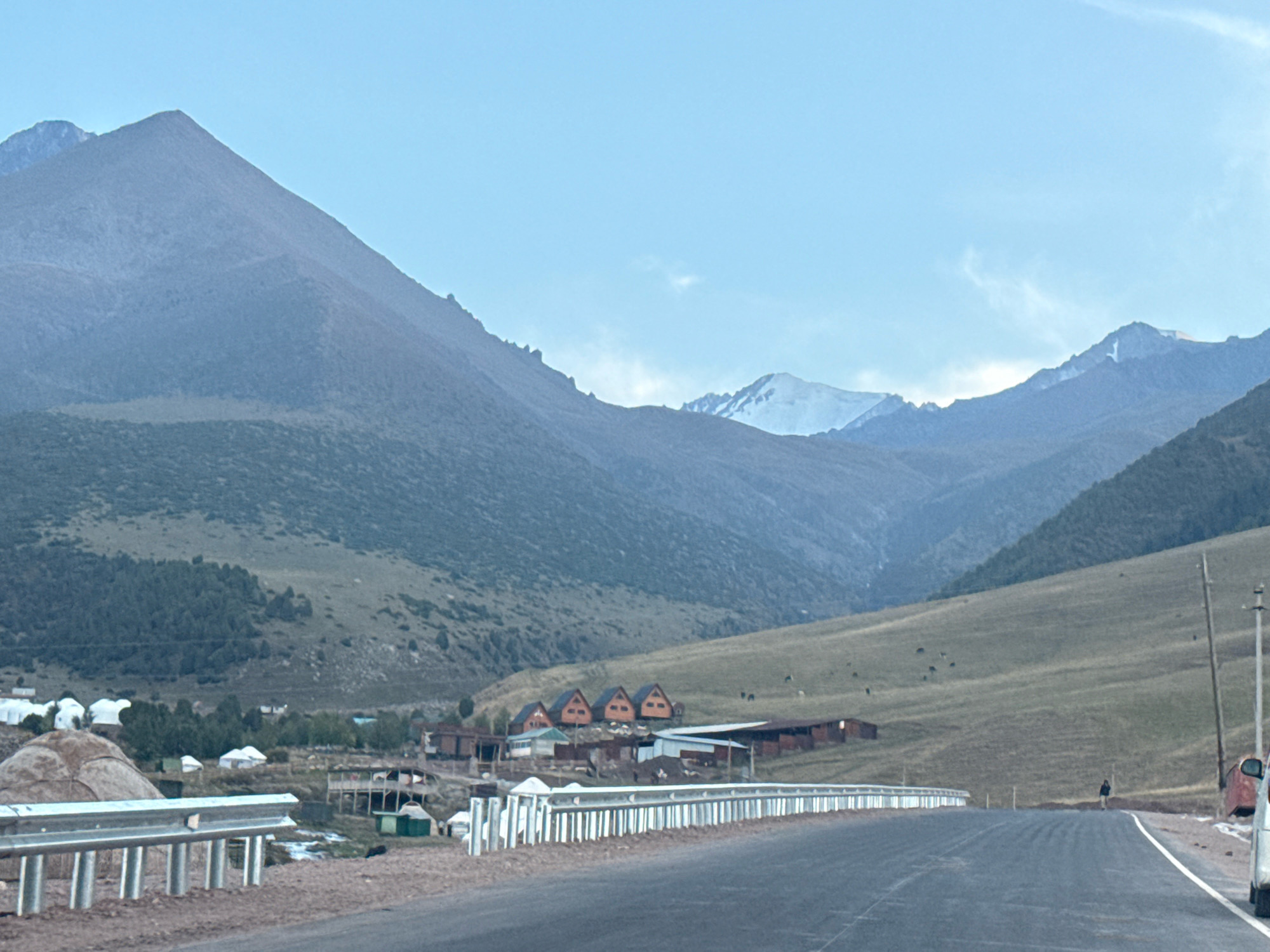 The image depicts a scenic mountain landscape in the city of Chunkurchak, Kyrgyzstan. A paved road curves through the foreground, bordered by a metallic guardrail. Beyond the road, a hillside is dotted with several small, A-frame style cabins, appearing to be part of a rural settlement or resort. In the background, imposing mountains dominate the horizon, with some peaks showing patches of snow. The sky is a clear, pale blue with a few wispy clouds. Several yurts, traditional nomadic dwellings, are visible further into the landscape. A lone figure stands on the side of the road in the middle distance. A white vehicle is partially visible on the right edge of the frame. The overall scene conveys a sense of remote, natural beauty and a peaceful, possibly rustic, setting.