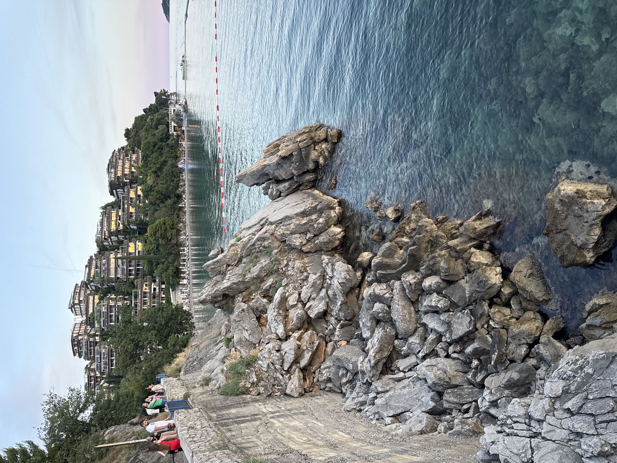 In Budva, Montenegro, a scenic view captures a rocky coastline leading to a tranquil body of water. On the left, a concrete retaining wall borders a pathway where several individuals are observed walking and standing. Among them are two women in red and black dresses, and a group of men and women. Behind them, a steep incline is covered with lush green trees, and terraced buildings with modern architecture are nestled into the hillside, their windows reflecting the soft twilight sky. The foreground is dominated by large, jagged rocks of various sizes that extend into the clear, blue-green water. A buouy line with red and white markers separates a swimming area from the open sea. In the distance, the horizon is marked by a faint landmass. The lighting suggests it is late afternoon or early evening, with a pale blue sky transitioning to a soft pink hue near the horizon.