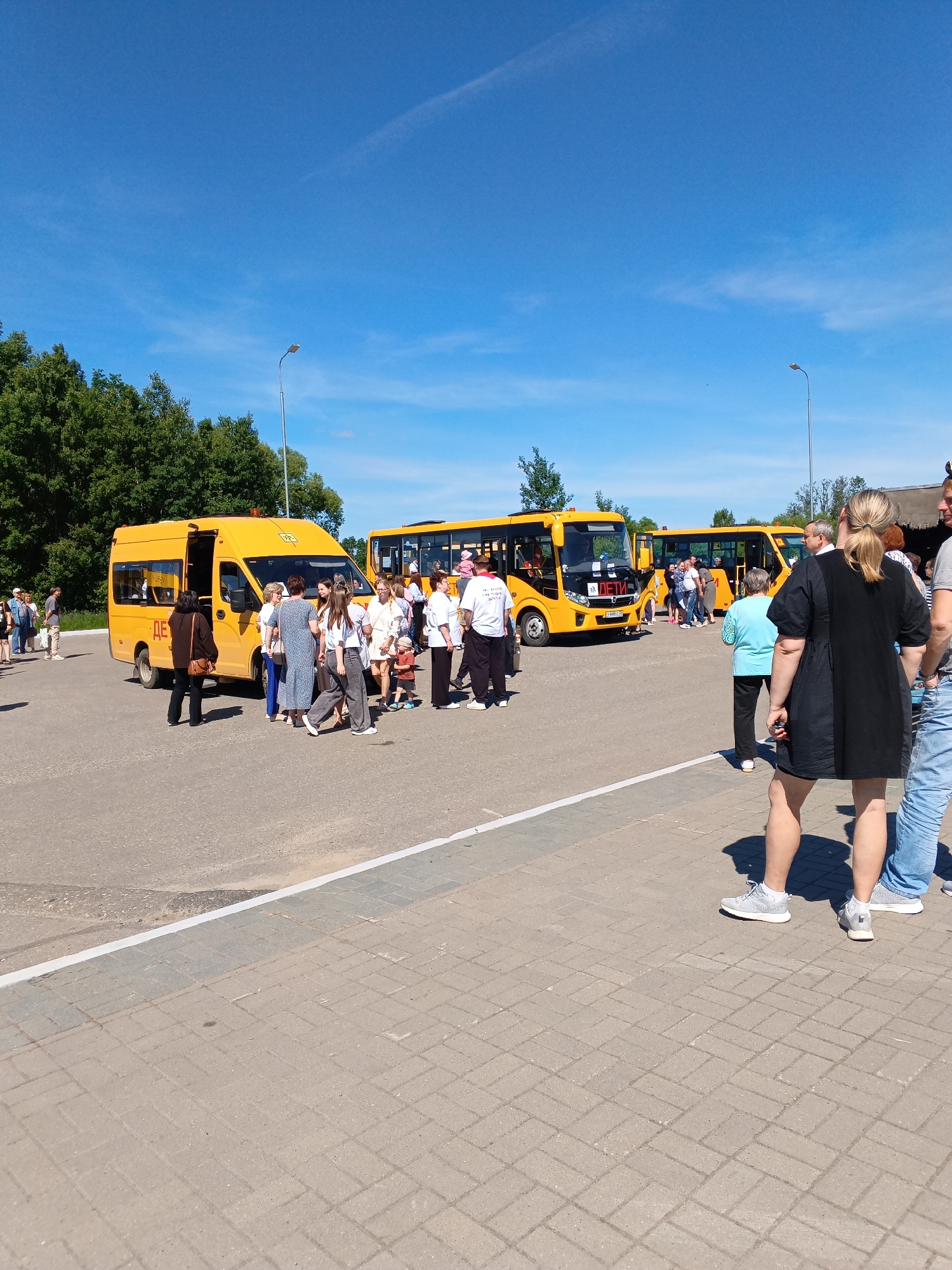 The image displays a bright, sunny outdoor scene in Sudogda, Russia, depicting a paved parking lot or gathering area. Three yellow vehicles, identified as school buses, are central to the composition. One smaller, van-style yellow bus is on the left, with its door open, prominently displaying the red Russian text "ДЕТИ" (CHILDREN) on its side. Two larger, conventional yellow school buses are parked further back towards the right, one of which also has the "ДЕТИ" text visible on its front.
Approximately 20-30 individuals, comprising a mix of adults and a few children, are congregated in groups around and between the buses. People appear to be engaged in conversations, waiting, or in the process of boarding or disembarking the vehicles. In the immediate right foreground, a woman in a black dress and white sneakers stands with her back partially to the viewer, next to a man whose lower body and arm are visible.
The environment includes a clear blue sky with minimal, wispy clouds. A dense line of green deciduous trees borders the background of the scene. A tall, silver street light pole stands in the mid-ground. The ground consists of grey asphalt for the vehicle area and beige rectangular concrete pavers for the foreground sidewalk, separated by a white-painted curb.