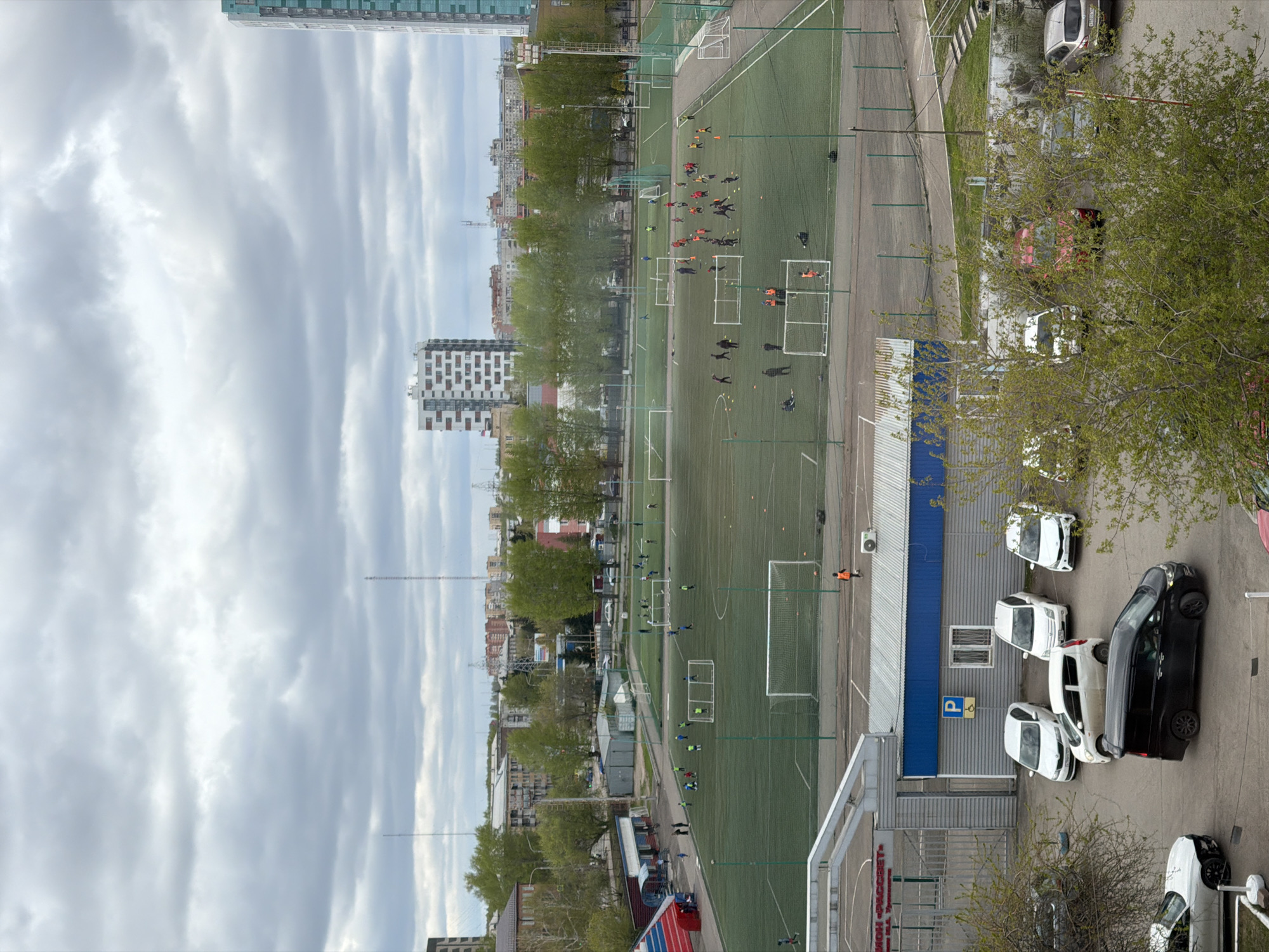An elevated, slightly off-center shot shows a soccer field located in what appears to be the city of Krasnoyarsk, Russia. The sky is overcast. People are scattered across the field, some appearing to be practicing or training. To the right of the field are several parked cars near a building. The surrounding area includes buildings and trees, typical of an urban setting.