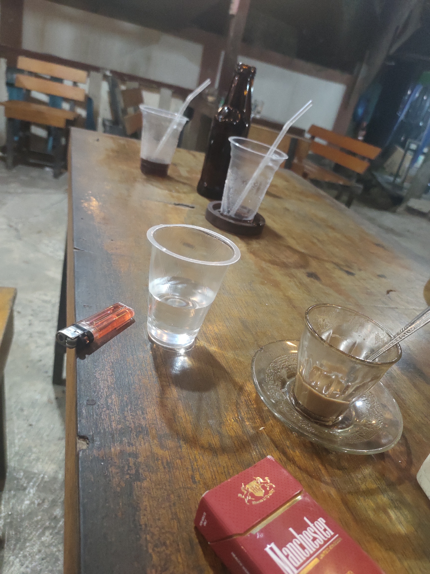 The image captures a dimly lit outdoor or semi-outdoor space in Keudematangkuli, Indonesia. A weathered wooden table is central to the composition. On the table, there are several drinking containers: two clear plastic cups with straws, one containing a dark-colored beverage, and the other appearing mostly empty. A brown glass bottle sits between them. A glass cup and saucer containing a milky coffee beverage with a spoon is also visible. Next to a plastic cup filled with water sits an orange lighter, and a red pack of "Manchester" brand cigarettes rests near the bottom edge of the frame. The environment suggests a casual gathering place, possibly a cafe or outdoor seating area. There is no evidence of people or activities in the image. The overall ambiance is subdued, indicative of a quiet evening or a low-traffic time of day.