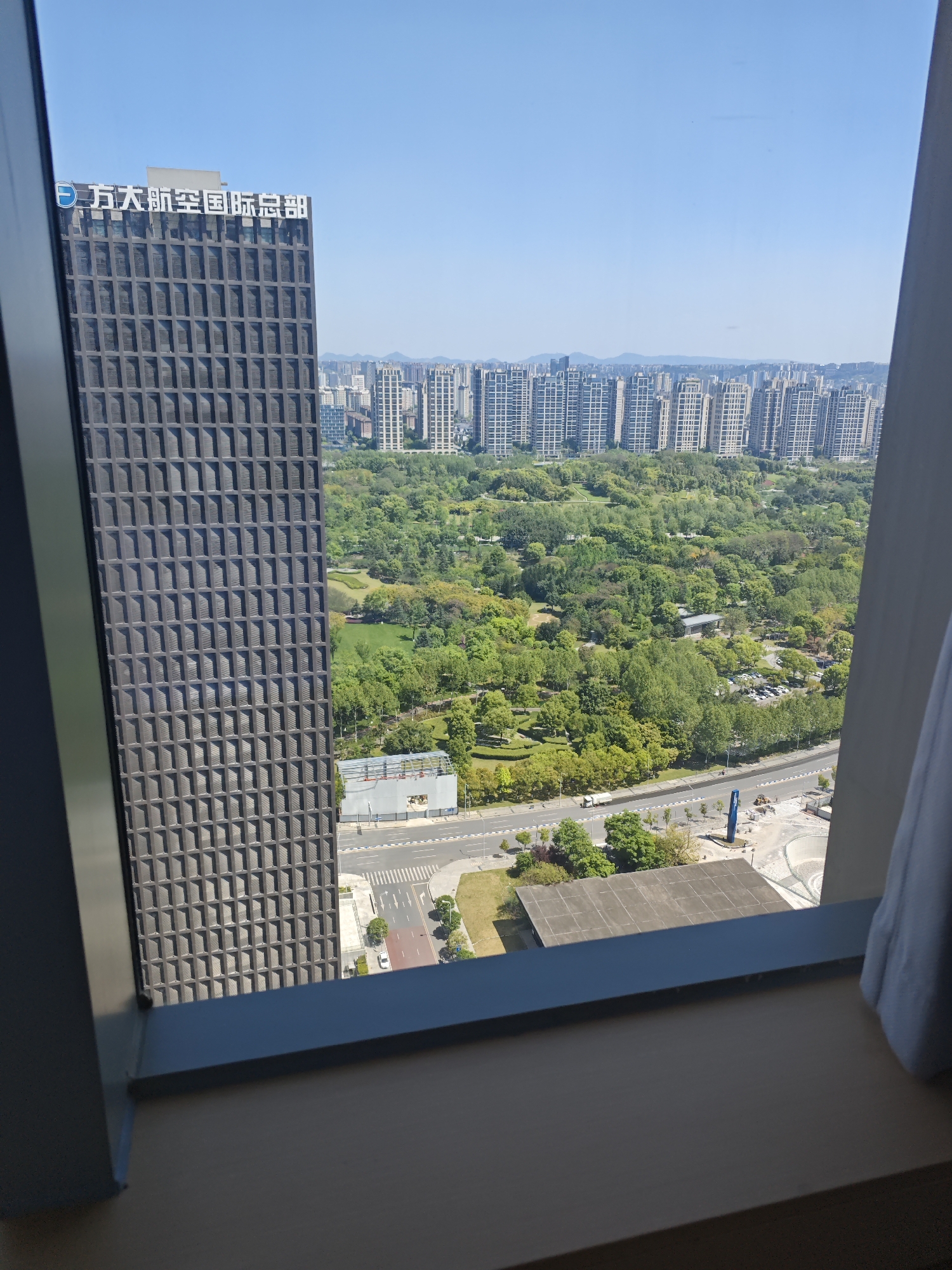 View from a window in Liangjiang, China. The image shows a dense urban landscape with a large park area, multiple high-rise apartment buildings in the distance, and a modern office building adjacent to the window. A two-lane road runs beside the park. Construction or landscaping activity is visible near the road. The sky is clear and blue.
