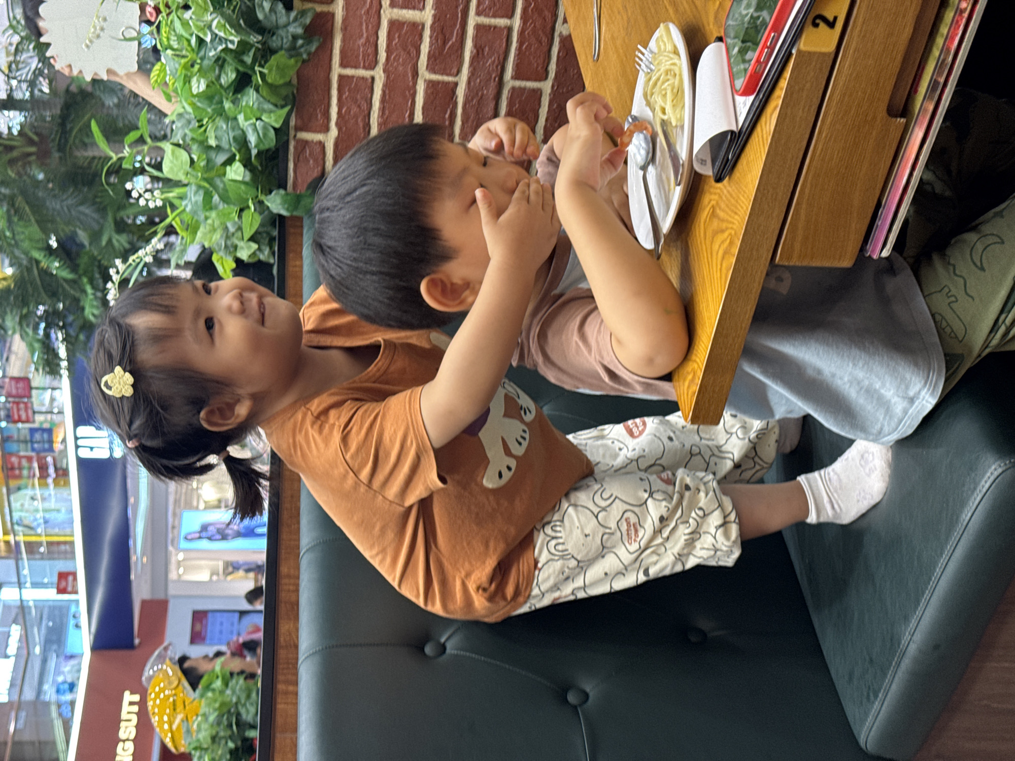 Two young children, a boy and a girl, are sitting at a table in what appears to be a restaurant. The table has a plate of spaghetti on it, along with cutlery and other items. The children seem to be interacting with the food, possibly eating. The girl is wearing a light brown shirt with a cartoon illustration. The background shows elements of a shopping mall or retail environment, visible through windows or openings. The location is indicated to be Haizhu District, China. The environment includes a booth with dark gray seating.