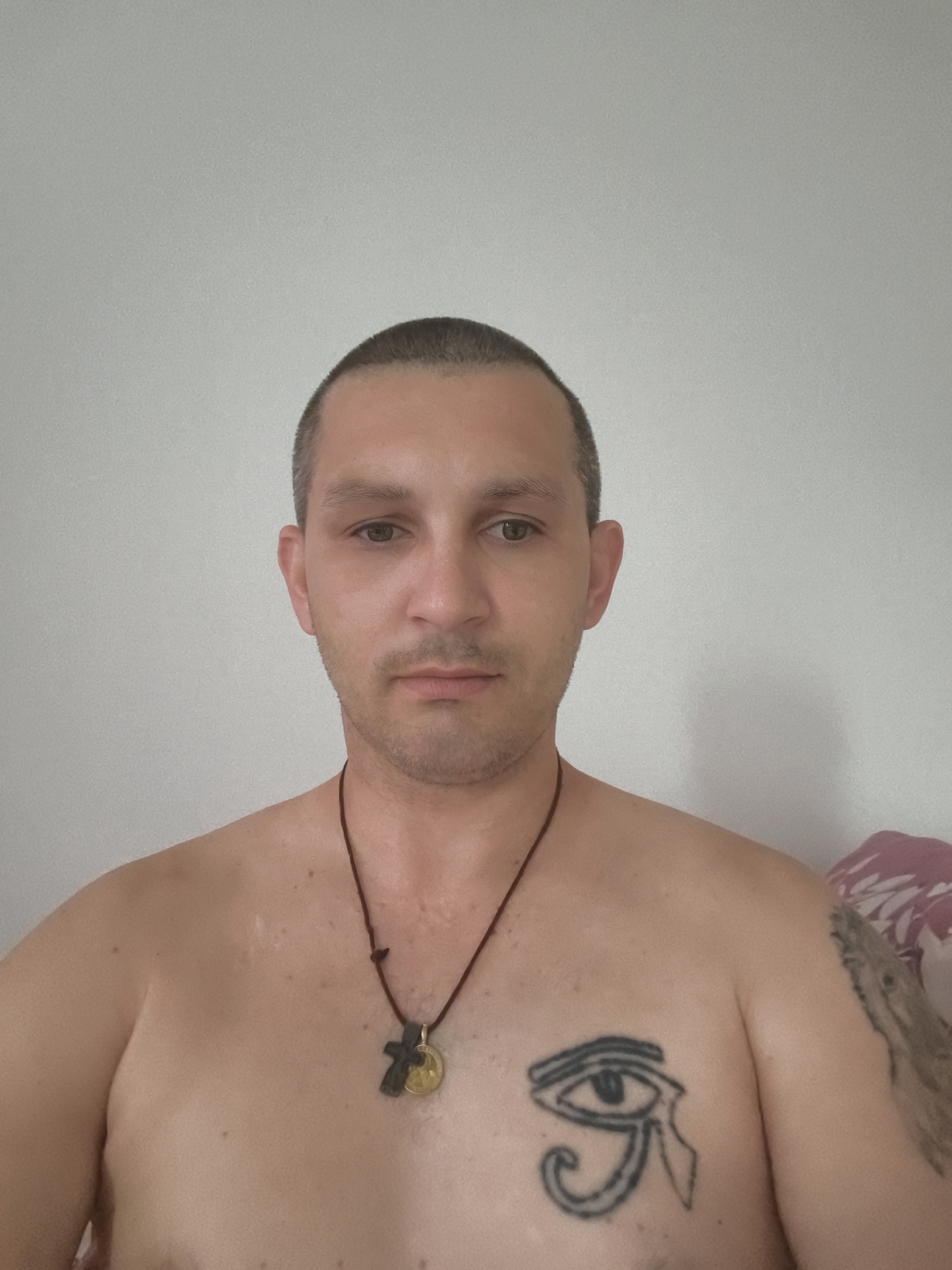 The image is a medium shot of a shirtless man with short, dark hair and a light complexion. He has a faint scar on his chest and a tattoo of the Eye of Horus on his left pectoral muscle. He is also wearing a necklace with a cross and a circular pendant. The man is looking directly at the camera. The background is a plain, light-colored wall. There is a hint of a patterned fabric on the right side of the image, possibly a pillow or blanket. The man is indoors, likely in a residential setting. The image does not contain any identifiable landmarks or features that would allow for the determination of a specific location within Wolgast, Germany.